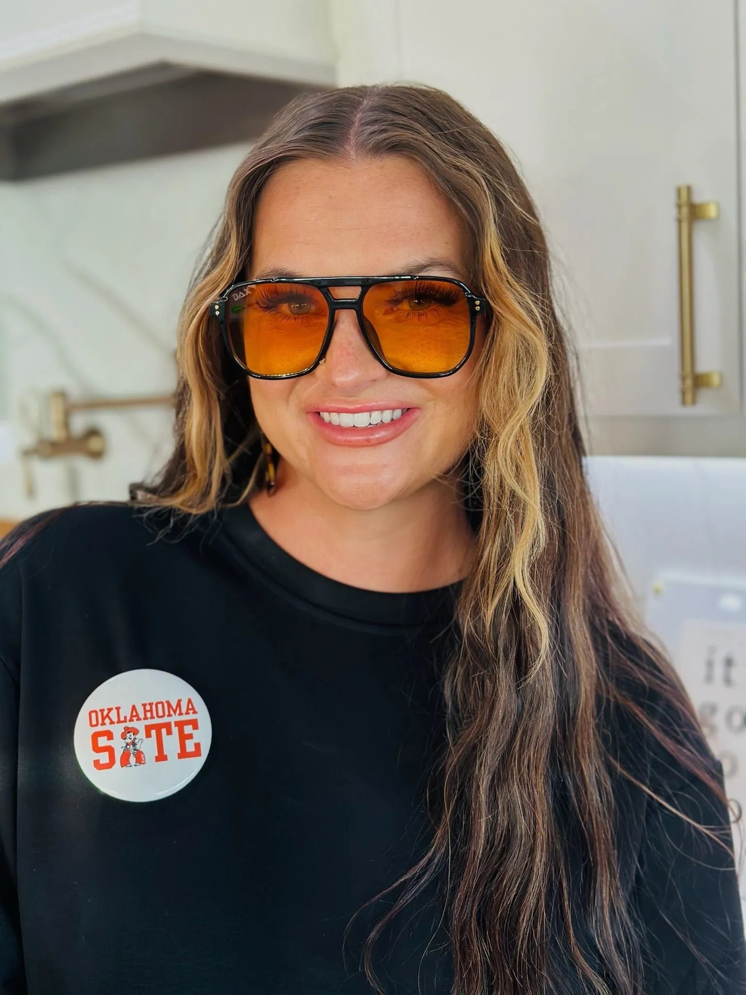 Woman wearing oversized sunglasses with orange lenses and a black shirt with an "Oklahoma State" button featuring the mascot.