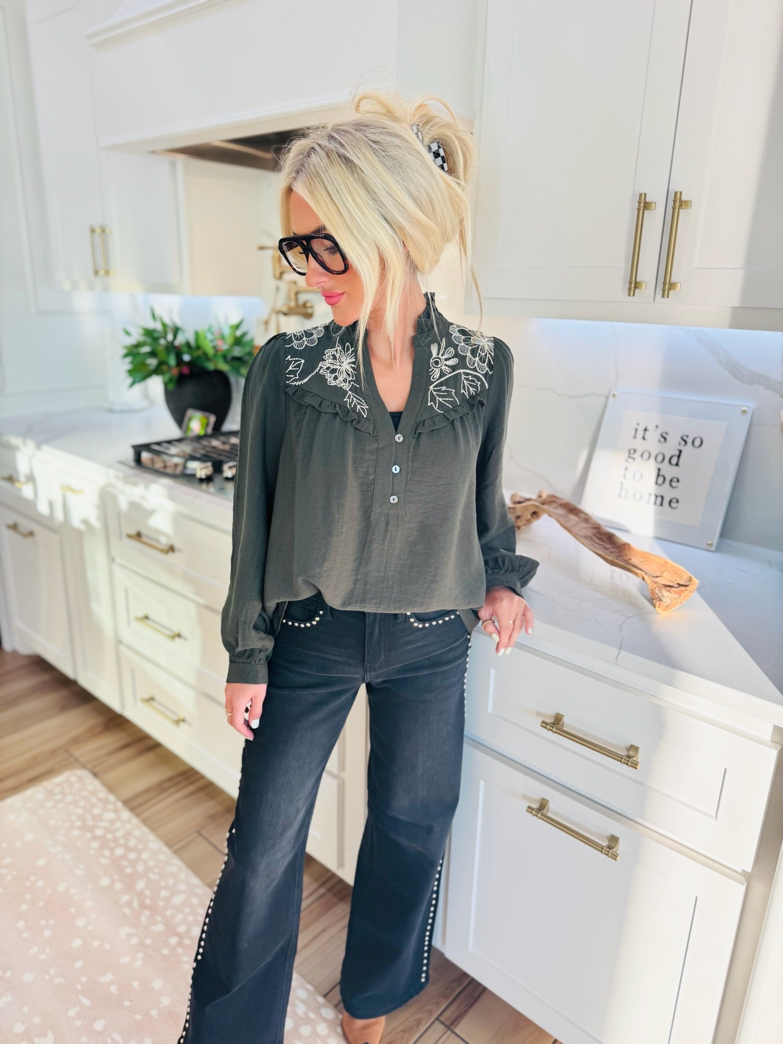 Olive floral embroidered blouse with ruffled collar and button placket, worn by a woman with blonde hair and glasses in a