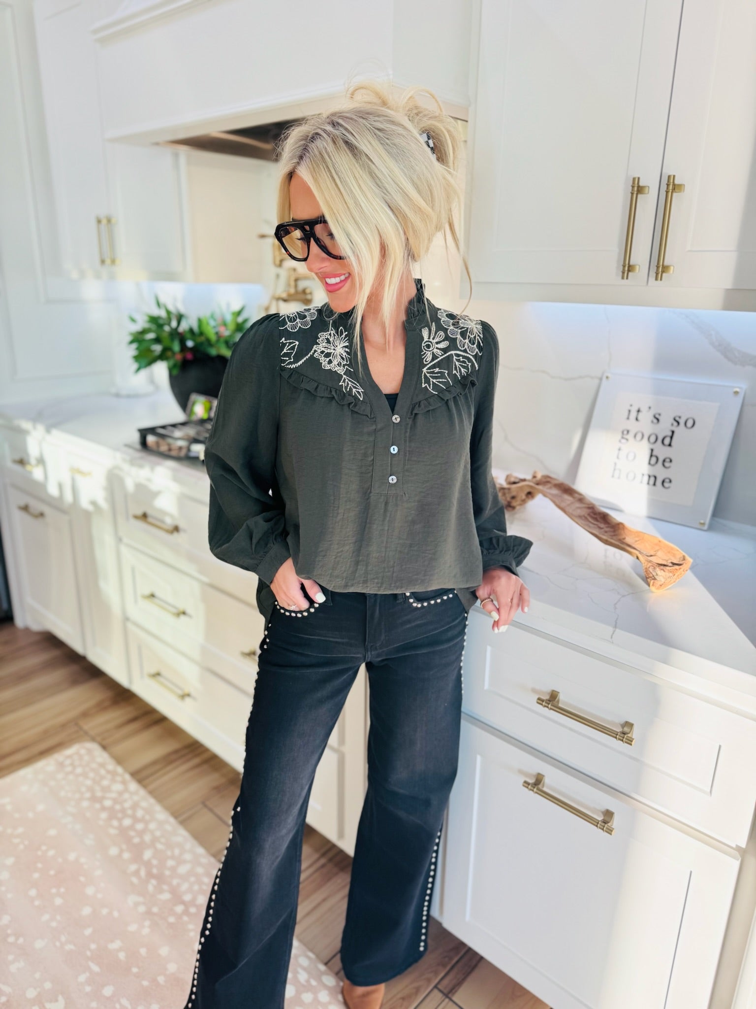 Woman wearing a dark olive green blouse with white floral embroidery and ruffled neckline, paired with dark wash flared