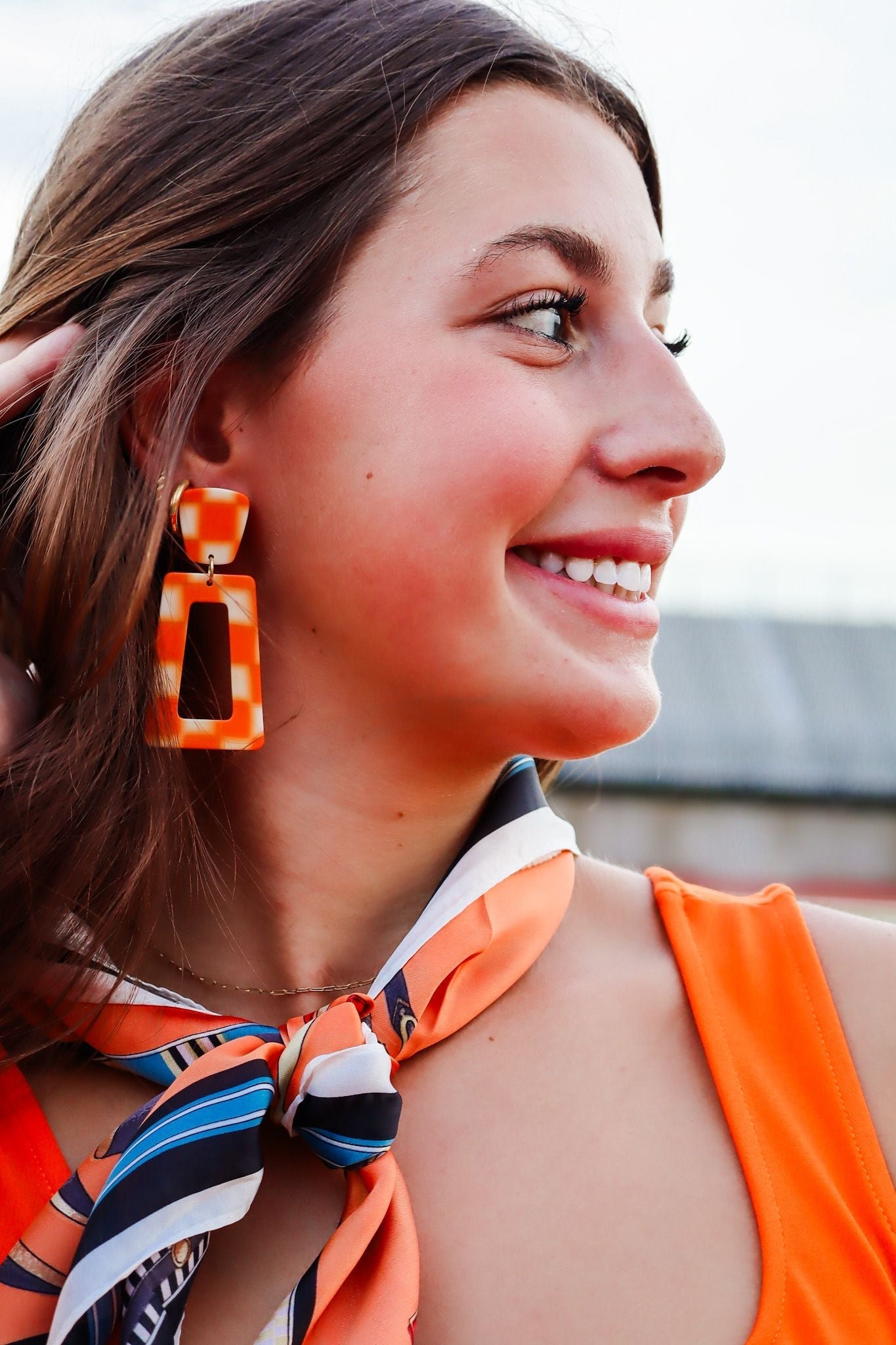 Orange and white checkered statement earrings with cut-out rectangular drop, worn by a smiling young woman.