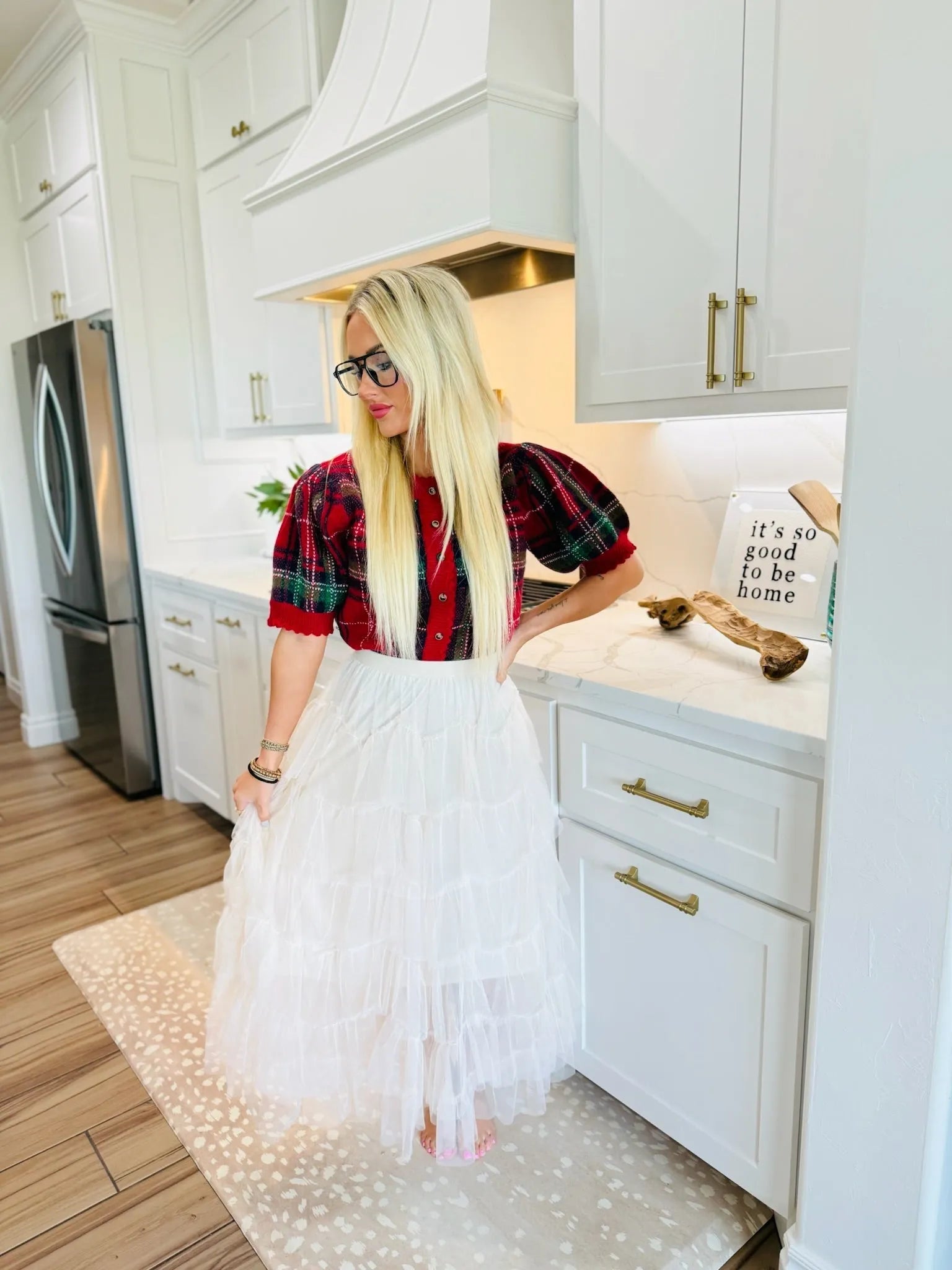 A blonde woman wears a white tiered ruffle mesh midi skirt and a red and green plaid puff-sleeve top. She stands in a