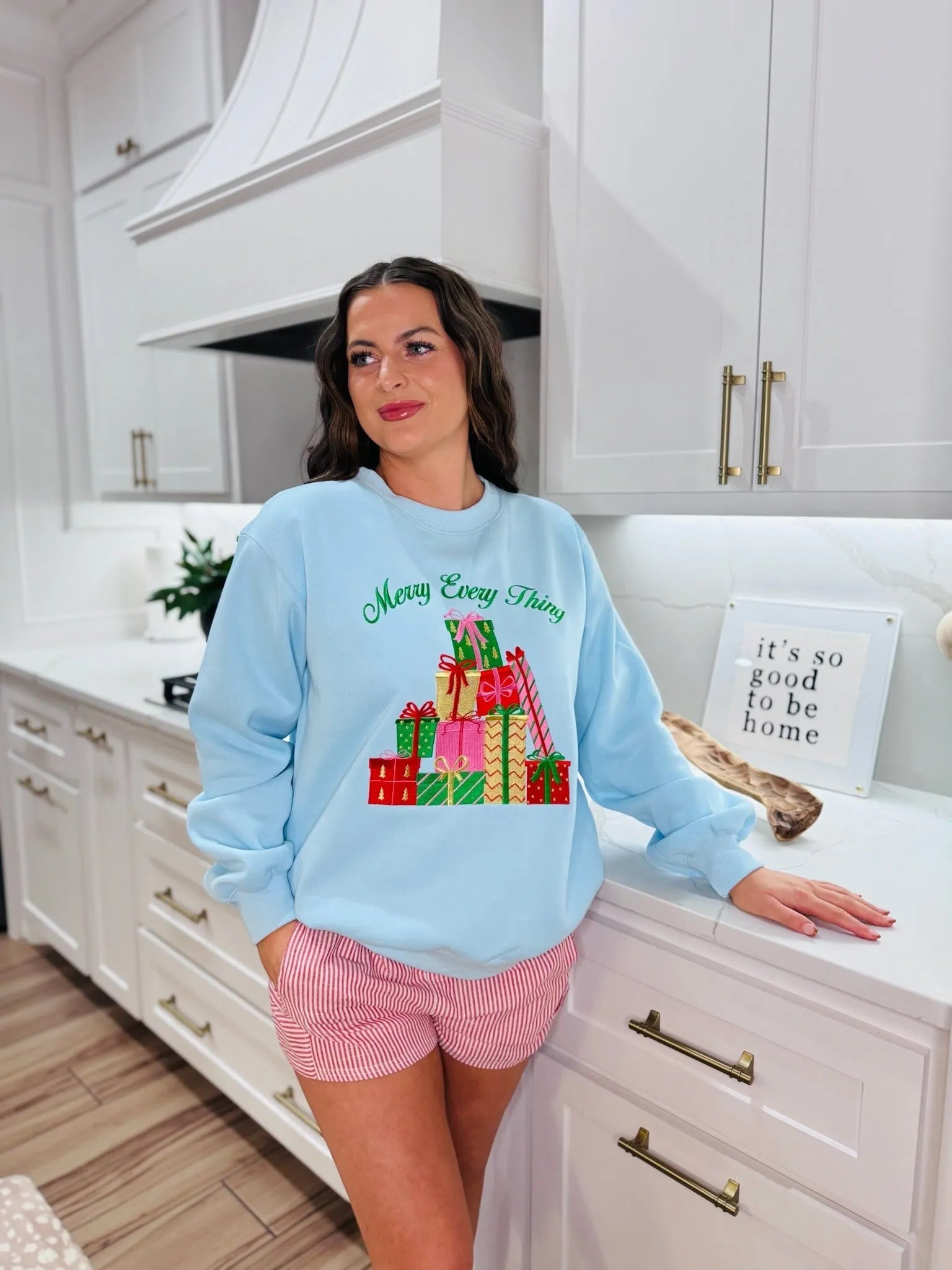 Light blue crewneck sweatshirt with green "Merry Every Thing" text and colorful stacked gift boxes graphic. Model wears red