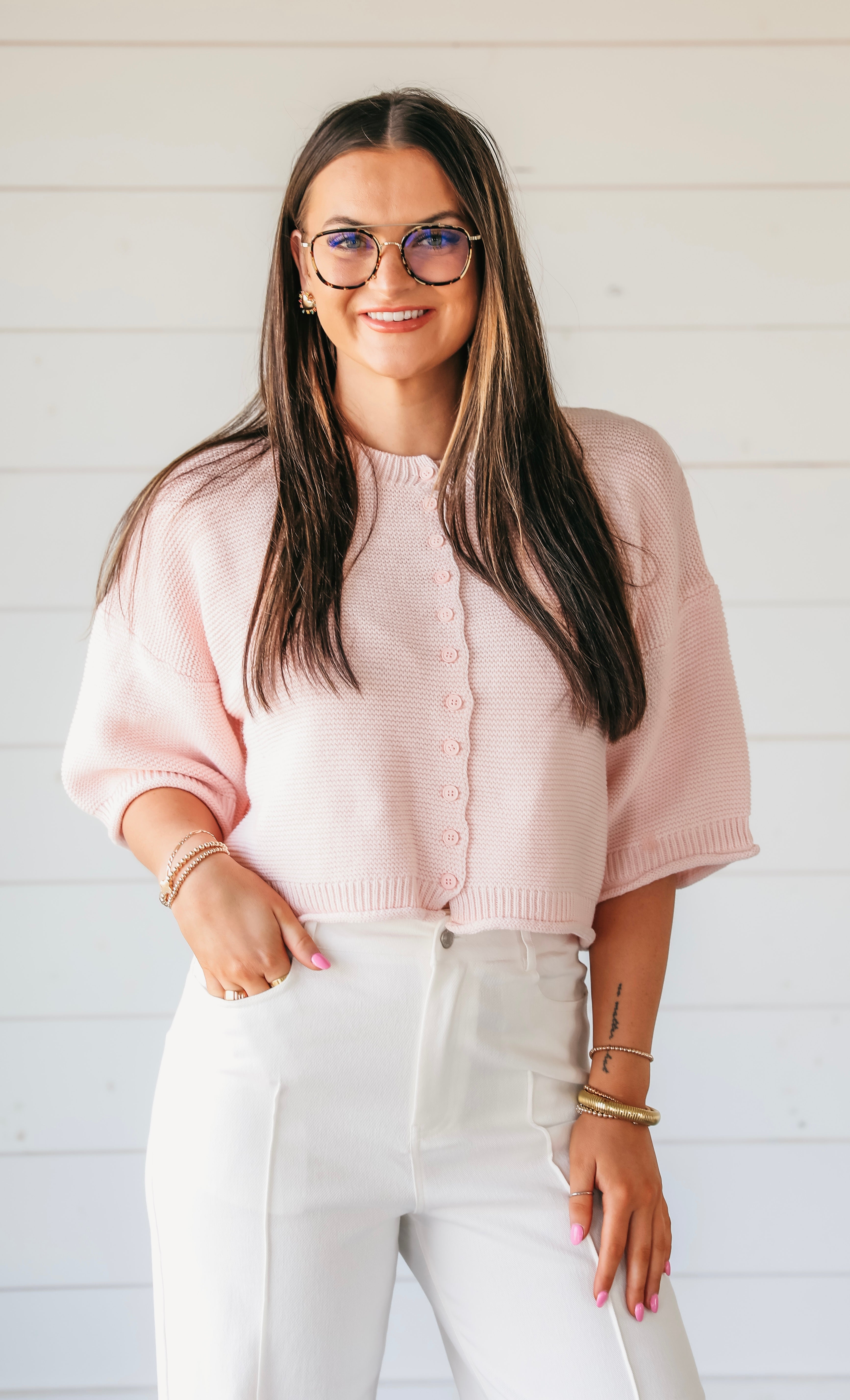 Woman wearing a pink waffle knit button-down cardigan sweater with cropped short sleeves and white pants.