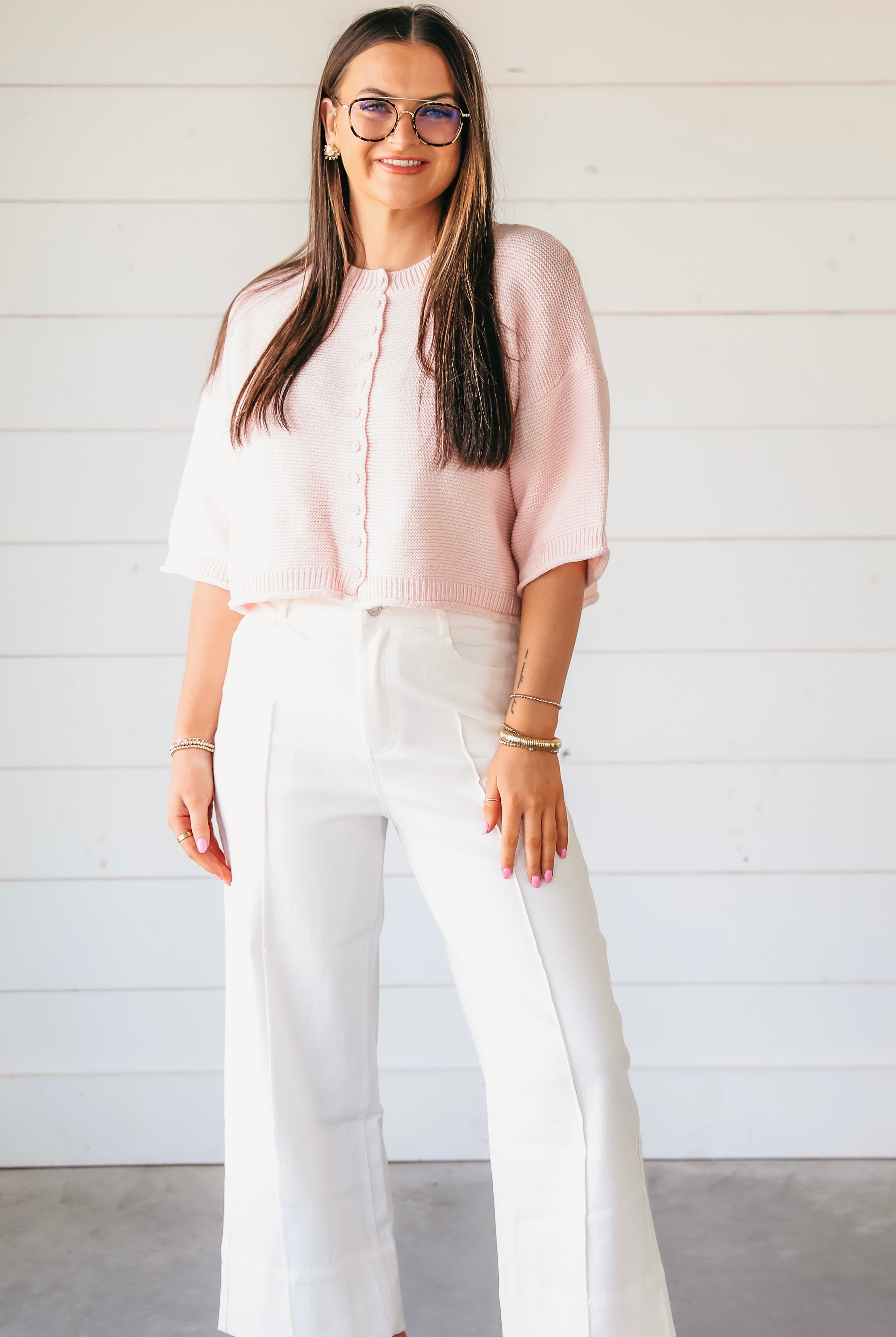 A woman wears a light pink waffle knit button-down cardigan sweater with white wide-leg pants.