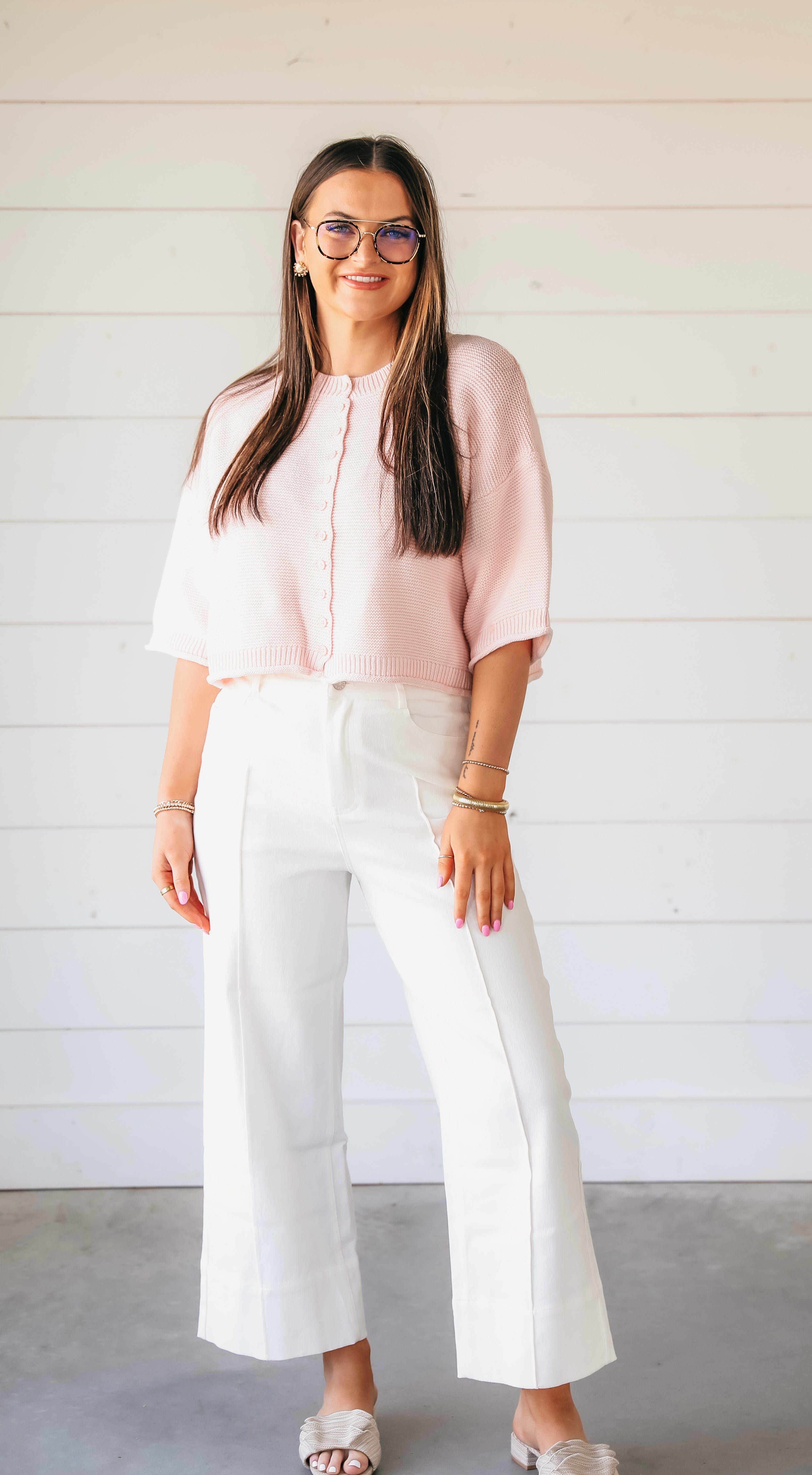 A woman wears a light pink waffle knit button-down cardigan sweater with white wide-leg pants.