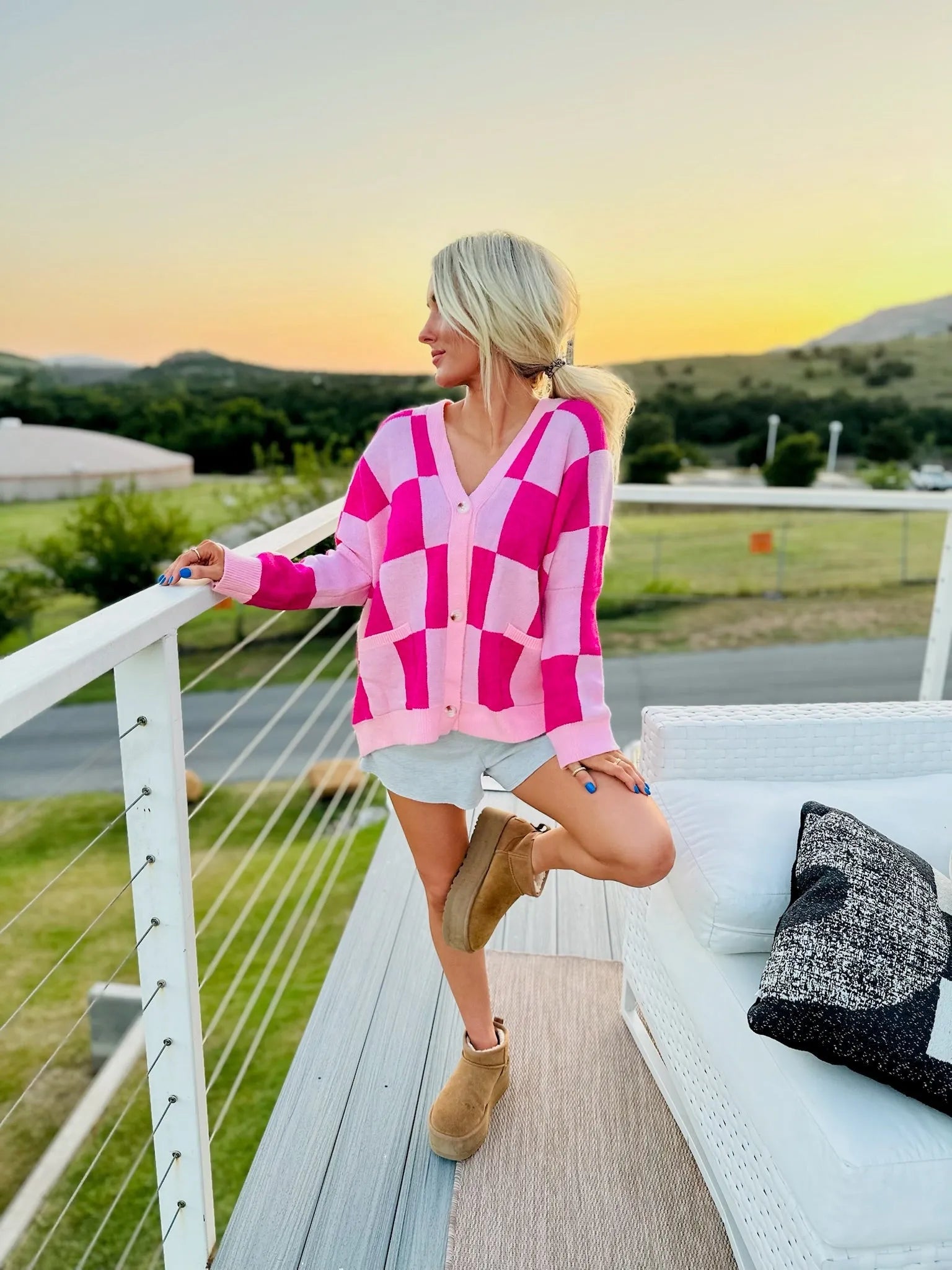 Woman wearing a pink checkered cardigan with a V-neck and button closure, paired with grey shorts and tan Ugg boots.