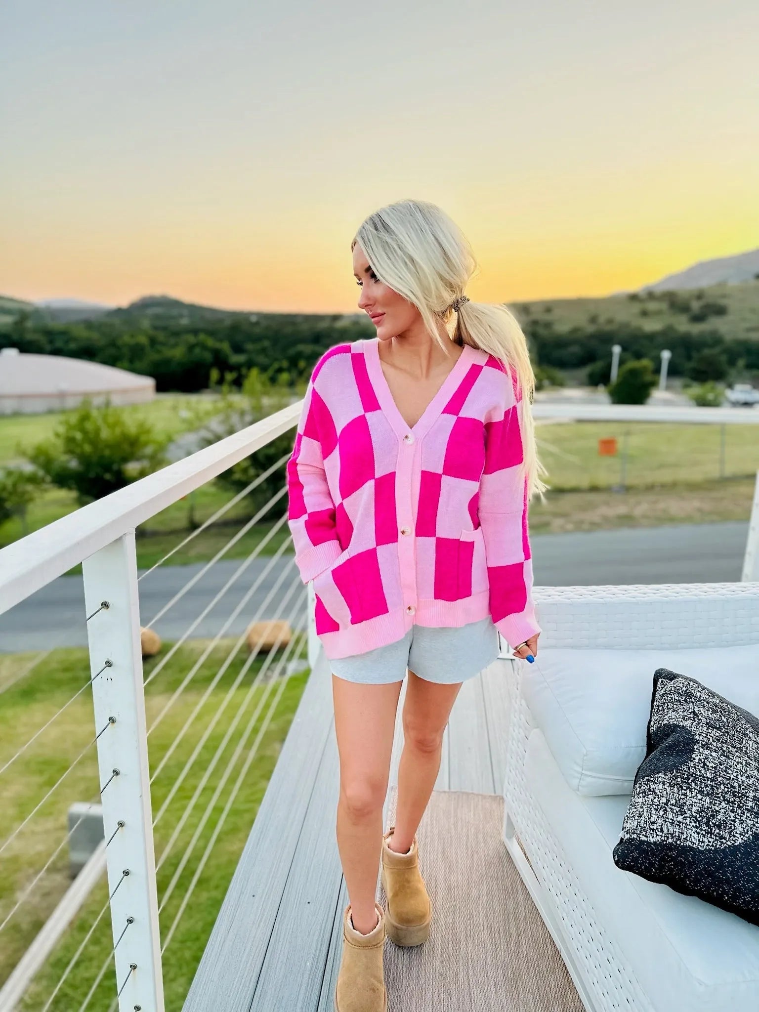 Woman wearing a pink checkered cardigan with button closure and pockets, light grey shorts, and tan boots.