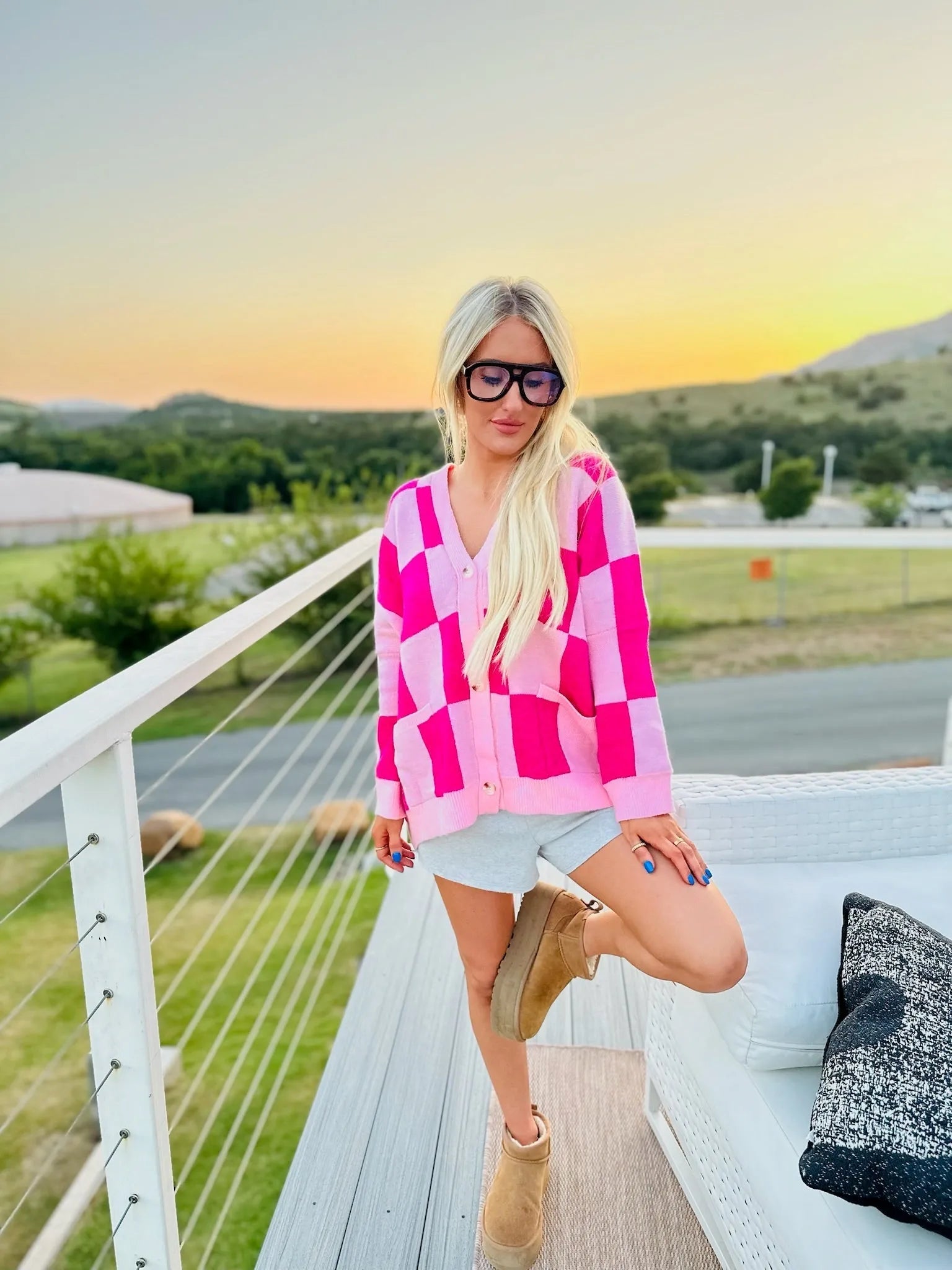 Woman wearing a pink and hot pink checkered cardigan, grey shorts, and tan Ugg-style boots.