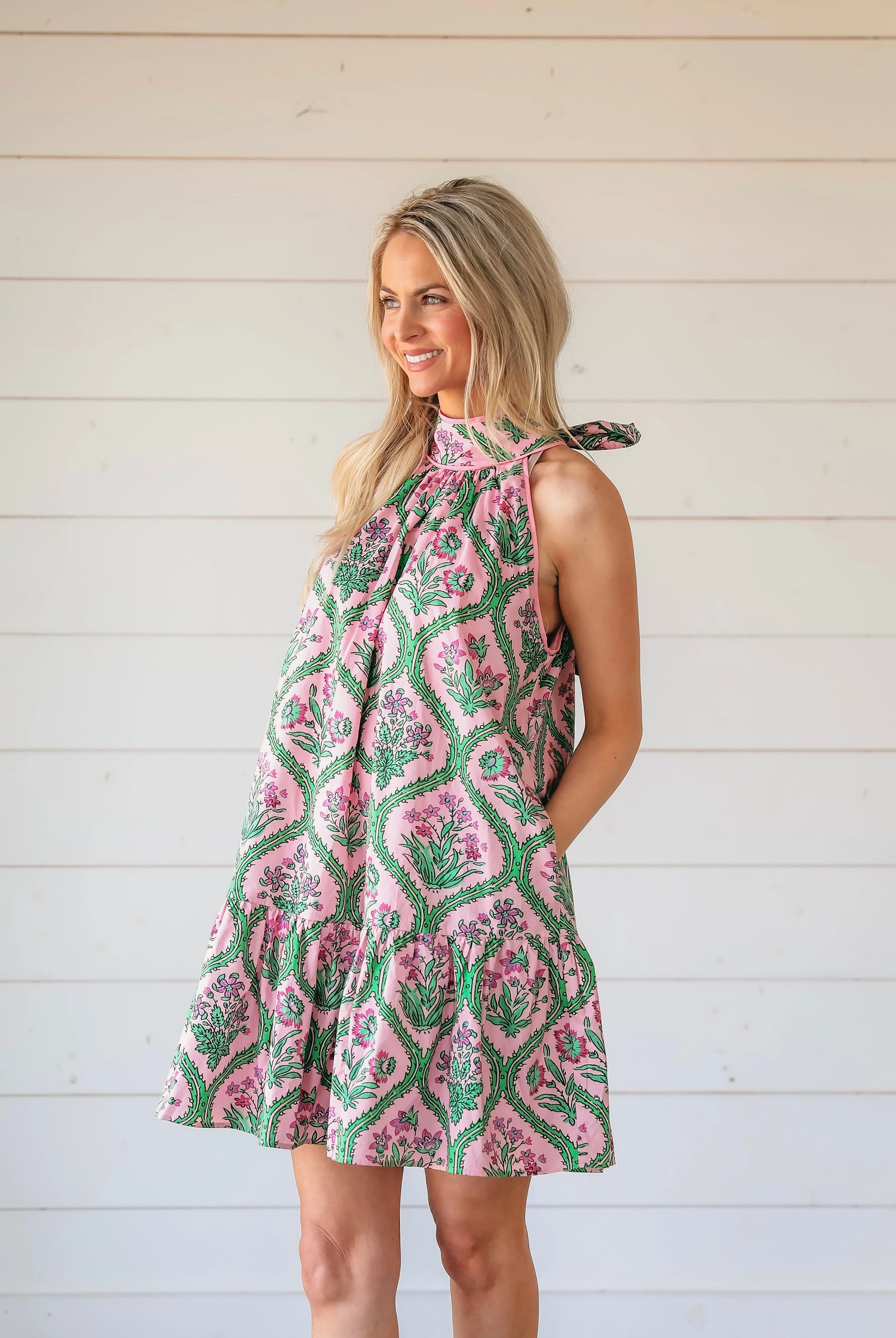 Pink floral halter mini dress with green vine pattern. Model smiling, hands in pockets. White wood background.