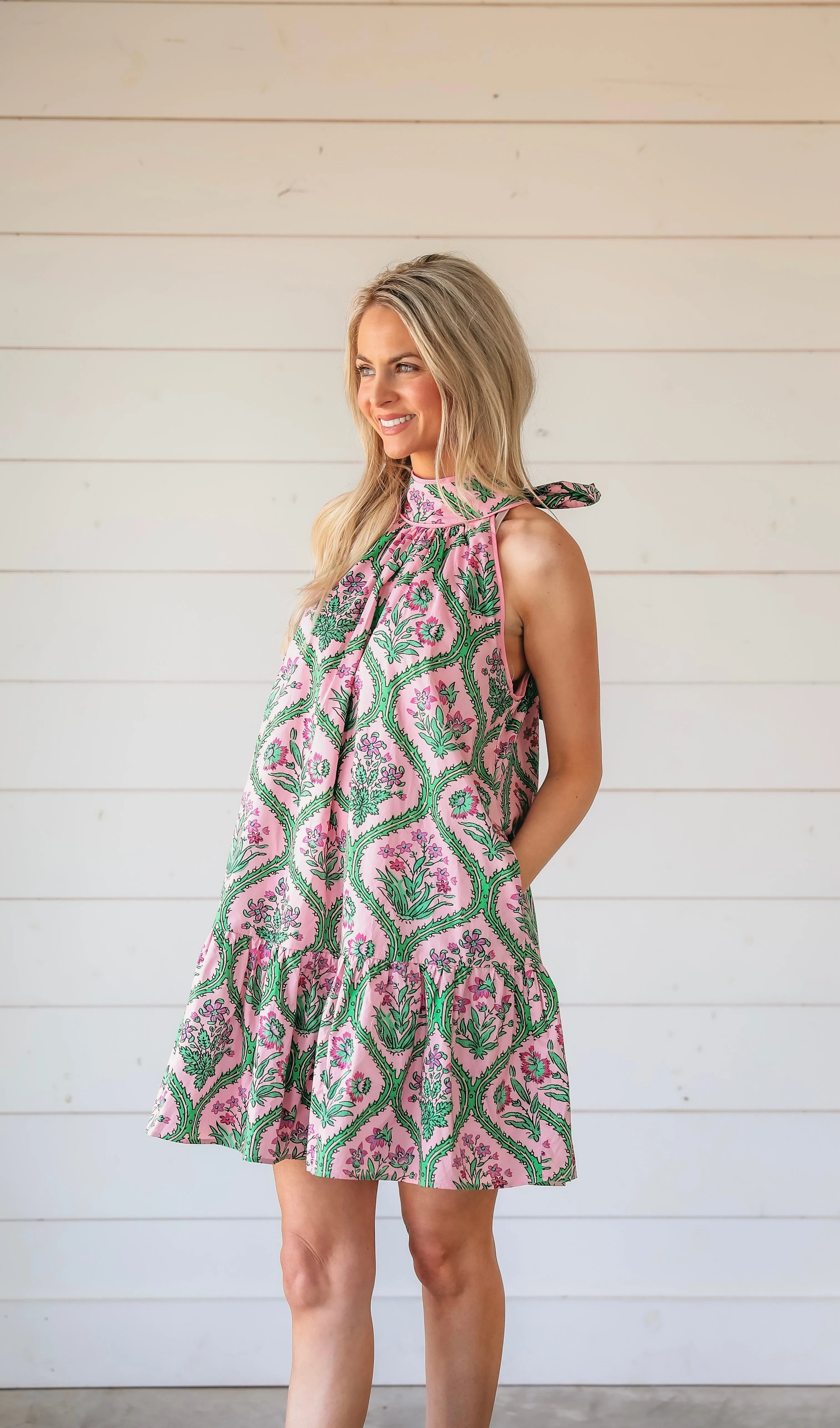 Pink floral halter mini dress with green vine pattern. Model smiling, hands in pockets. White wood background.