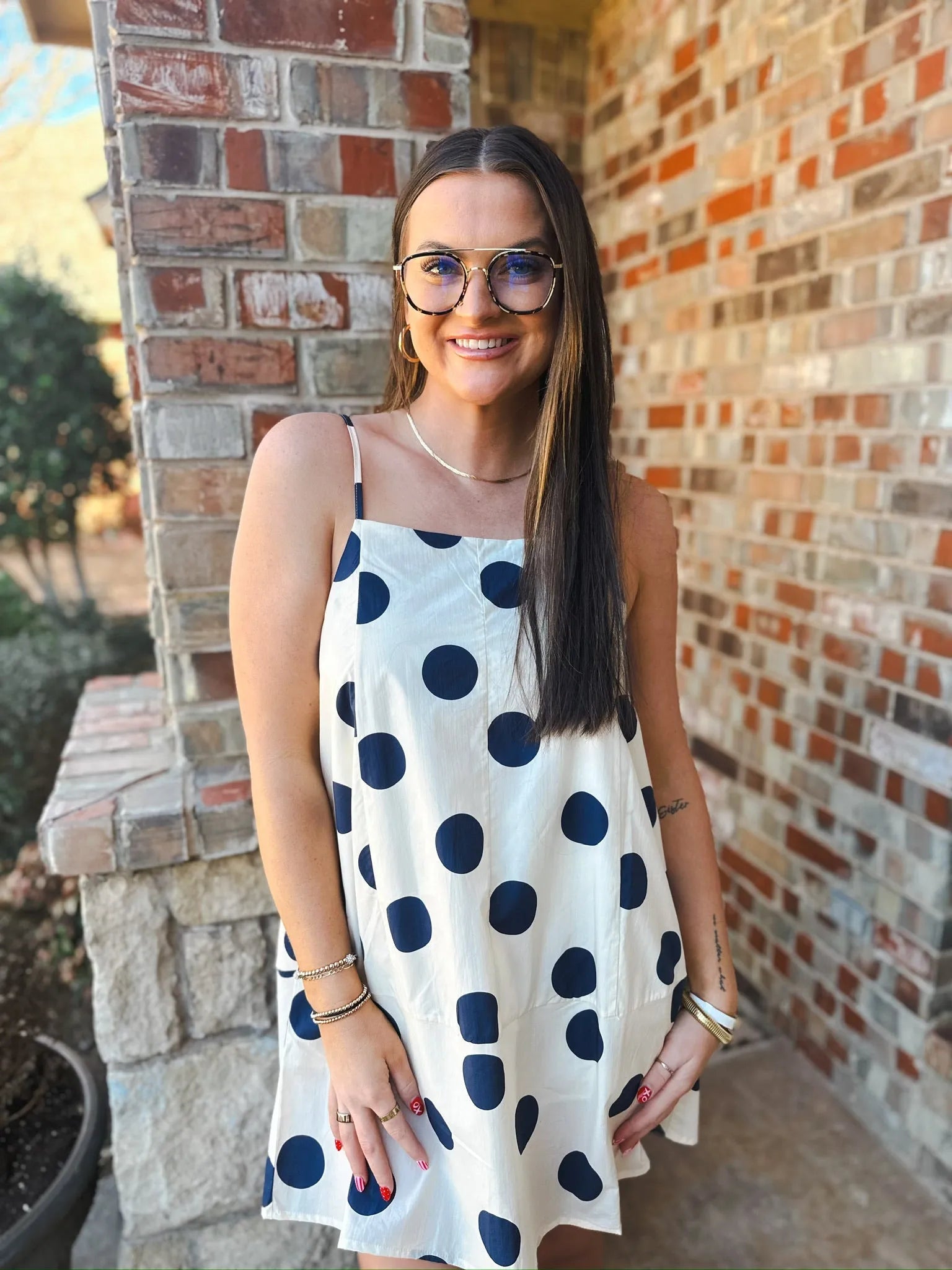Woman wearing a navy and cream polka dot mini dress with spaghetti straps, gold aviator glasses, and jewelry.