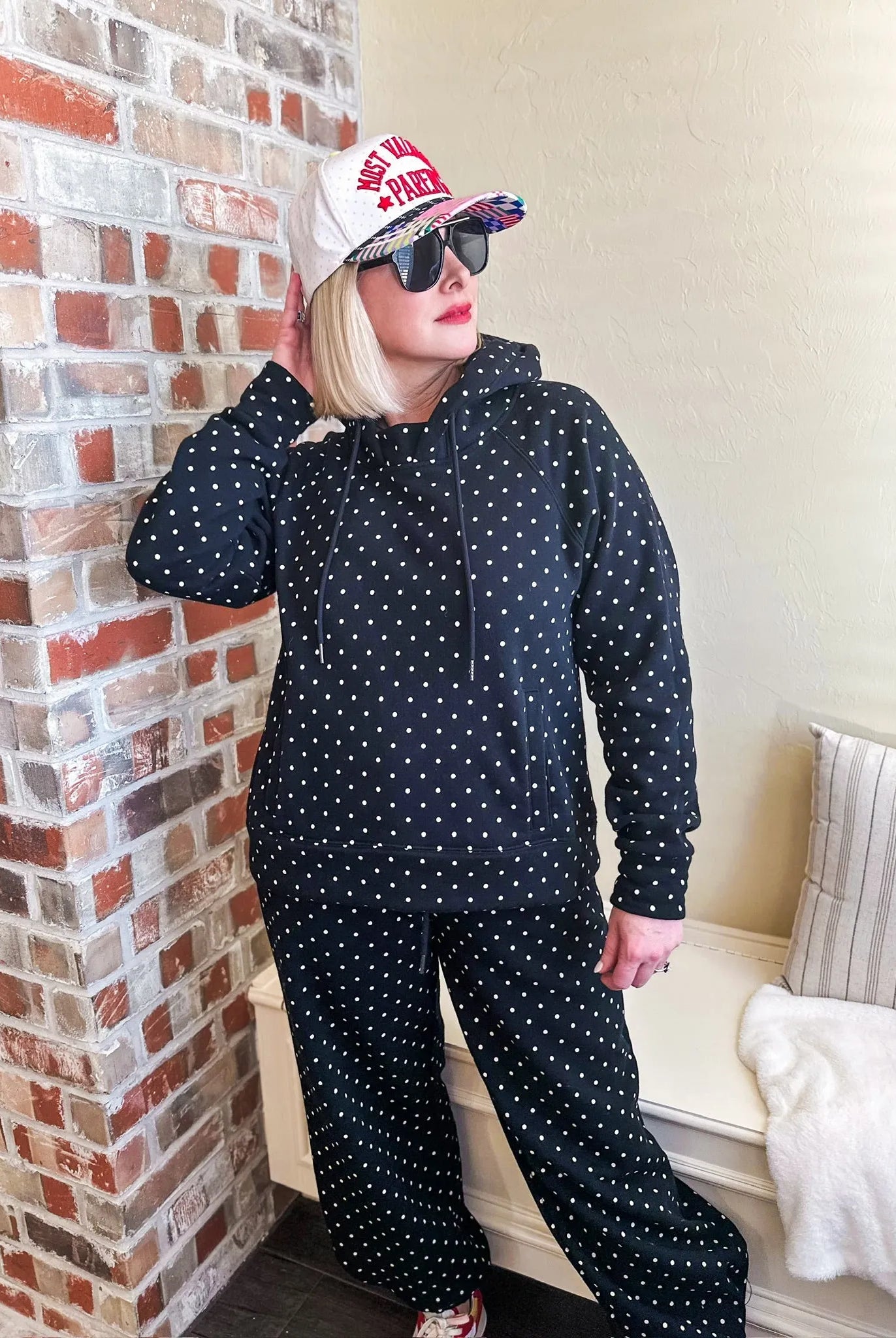 Black and white polka dot sweatsuit with drawstring hoodie and pants. Model wears a baseball cap and sunglasses.