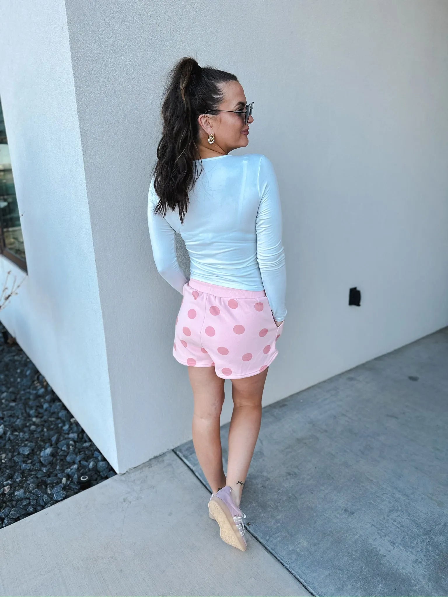Poppy & Pine blush pink polka dot shorts. Model from behind wearing white long-sleeve top, pink polka dot shorts, and pink