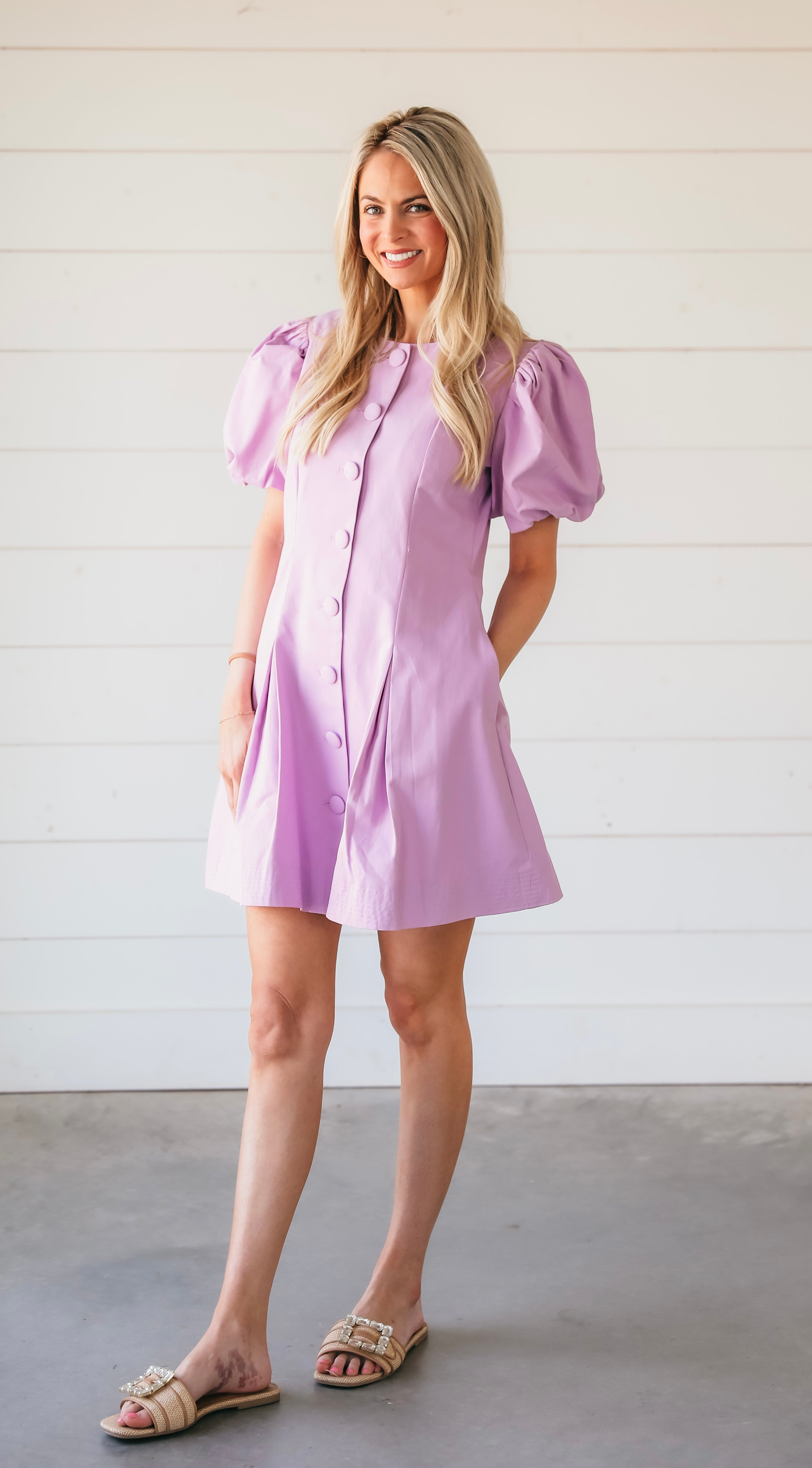 Woman wearing a purple puff sleeve structured mini dress with button front and pleats. She is standing with hands in pockets.