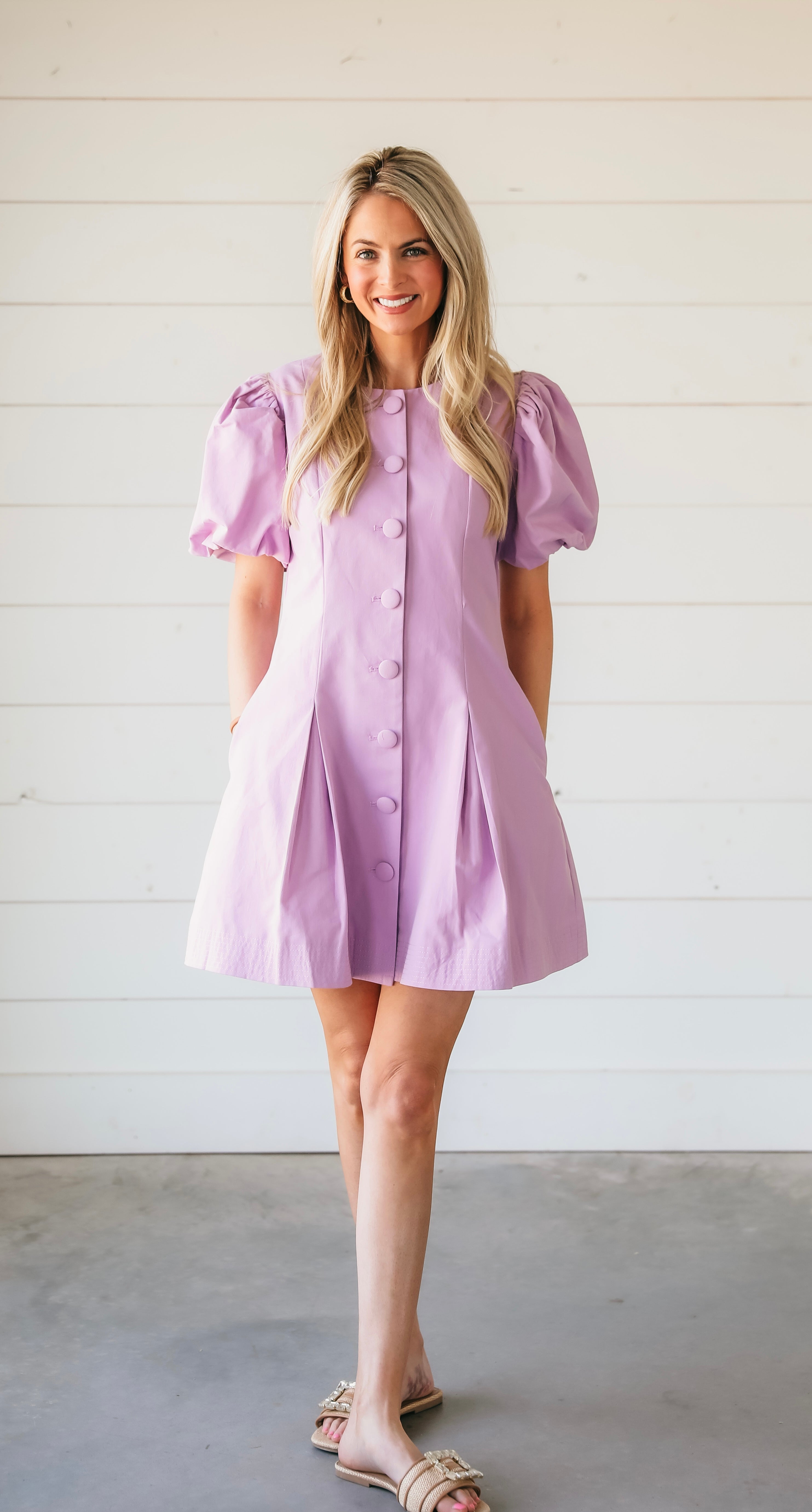 Woman in a structured purple mini dress with puff sleeves, button-down front, and pleated skirt. Stands against a white