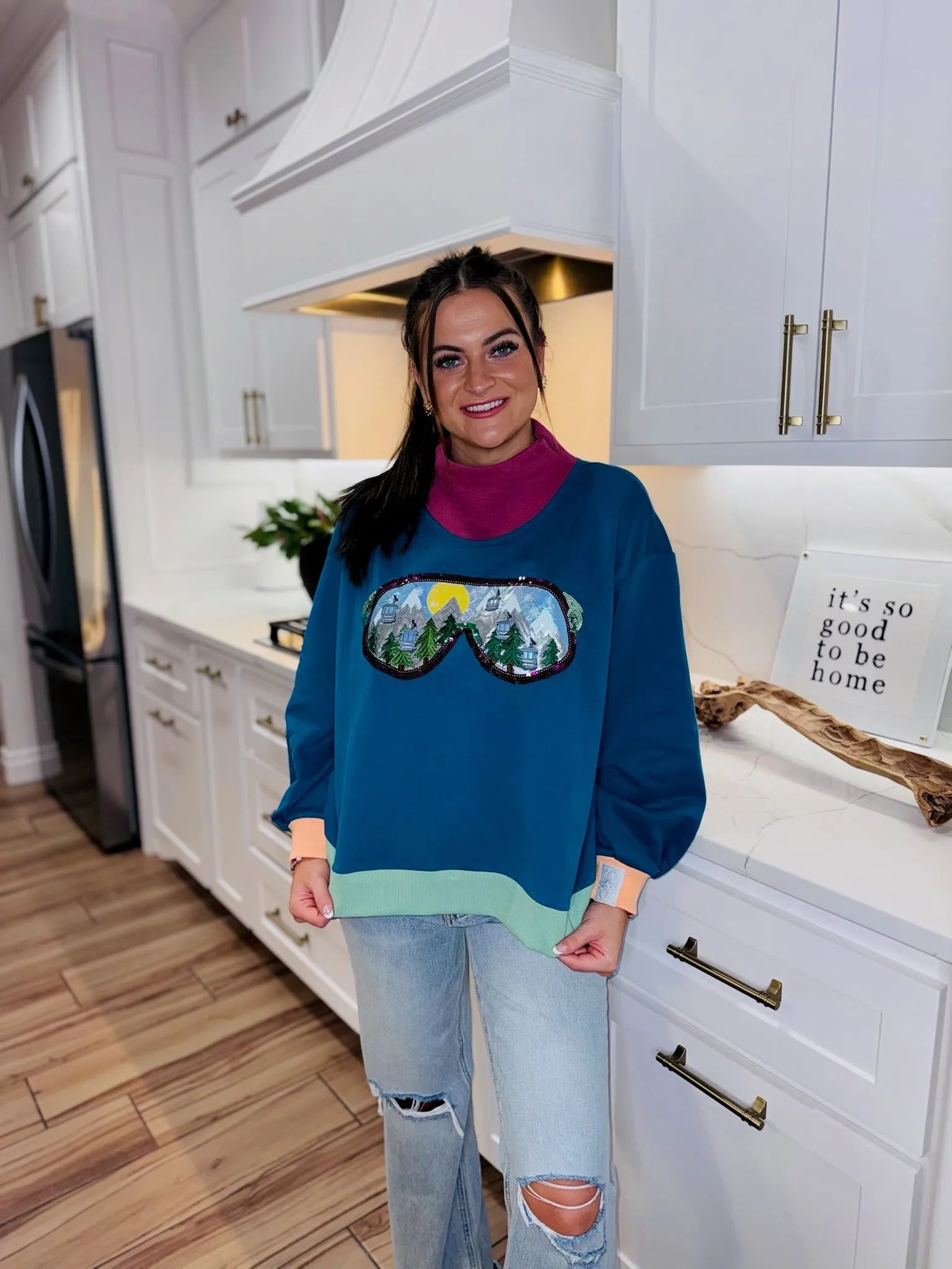 Woman wearing a blue sweatshirt with a ski goggle reflection graphic, featuring mountains, trees, and ski lifts. The