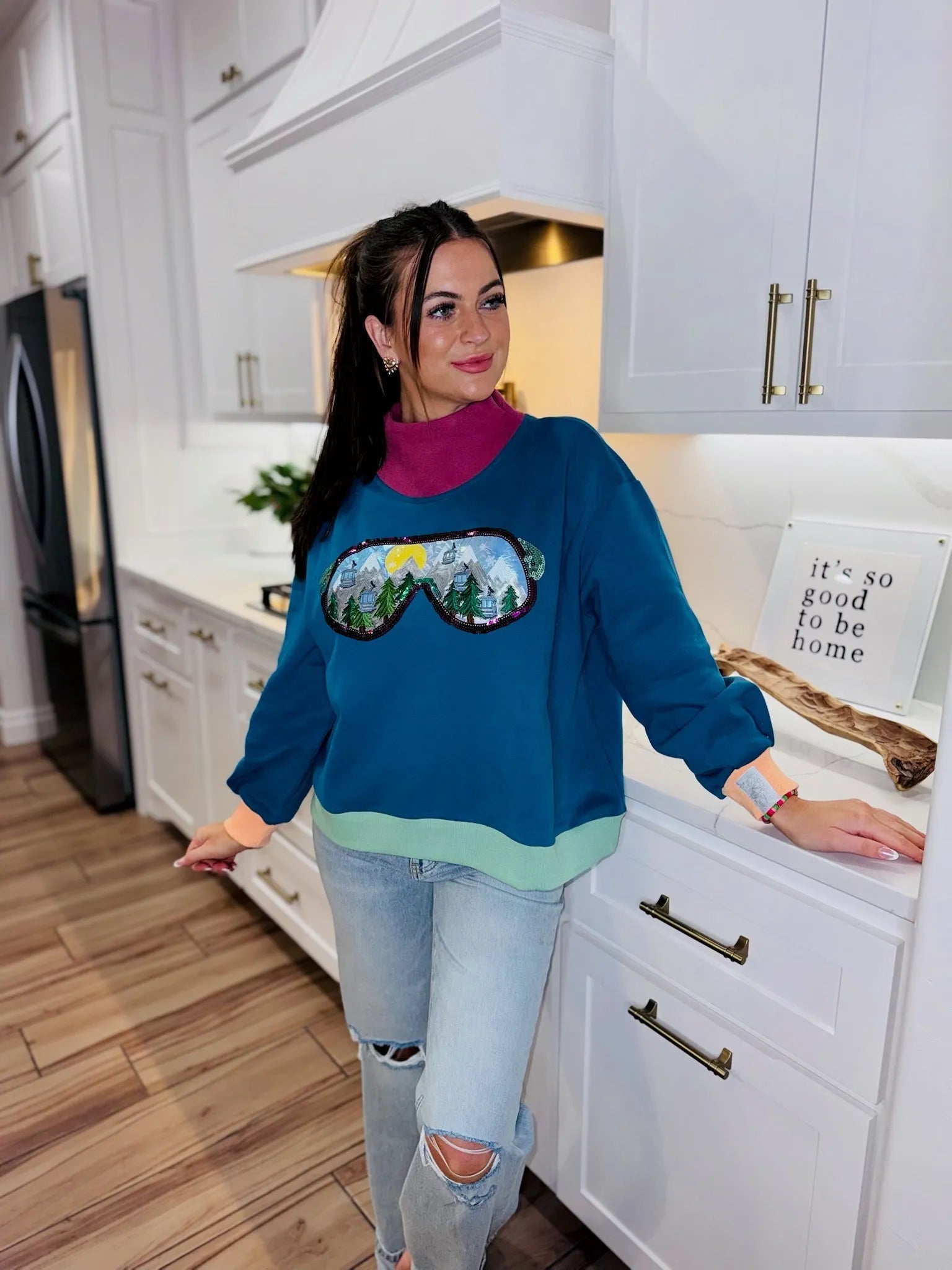 QOS blue sweatshirt with a ski goggle graphic featuring mountains, trees, and a yellow sun. Light green hem and pink