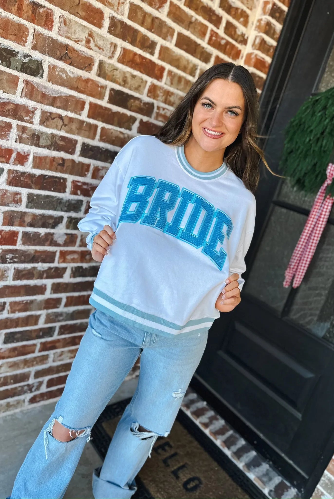 White long-sleeve sweatshirt with blue rhinestone "BRIDE" text and light blue stripes on cuffs and hem. Worn by a smiling