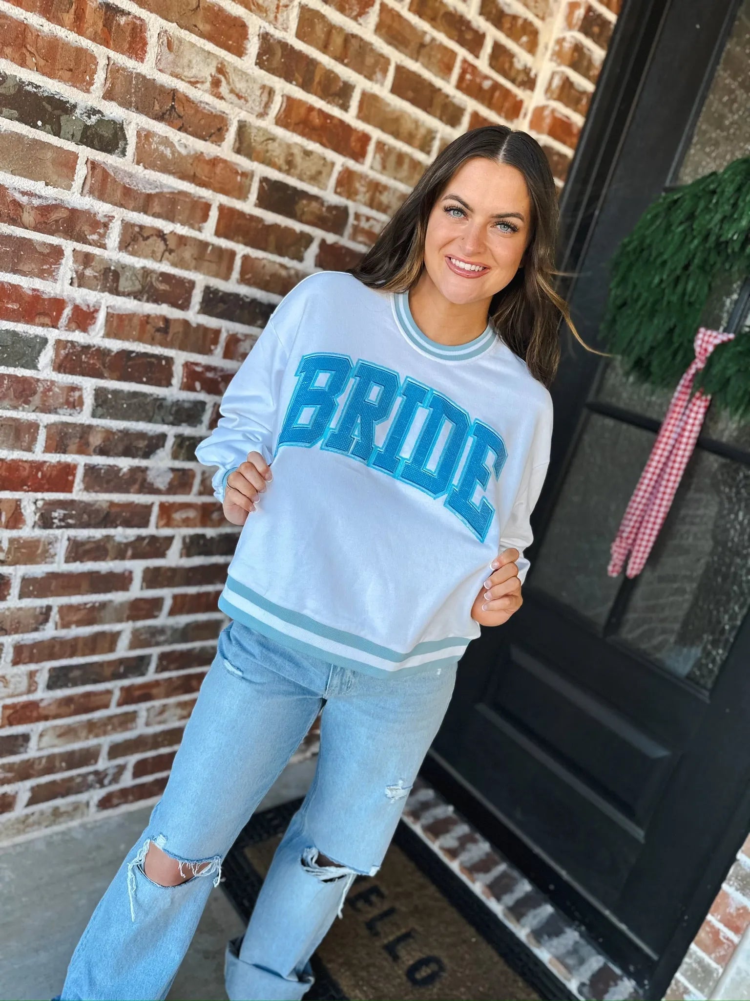 White long-sleeve sweatshirt with blue rhinestone "BRIDE" text and light blue stripes on cuffs and hem. Worn by a smiling