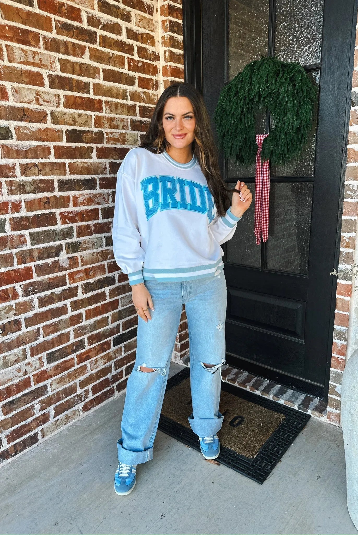 Woman wearing a white "Bride" sweatshirt with blue lettering and trim, distressed light wash jeans, and blue sneakers. She