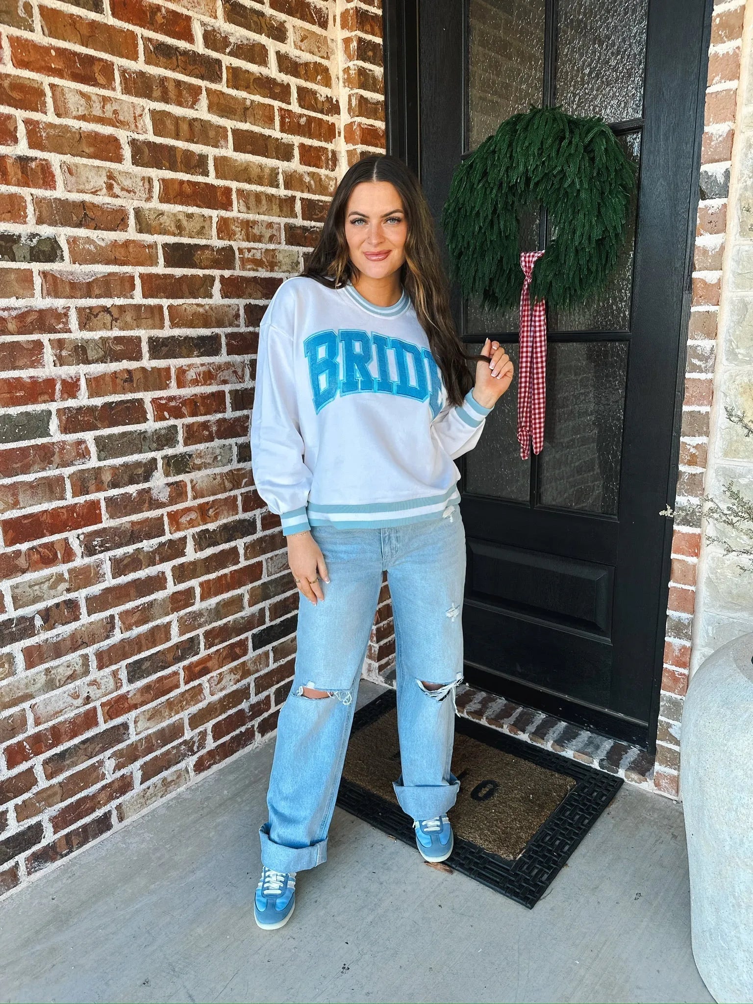 Woman wearing a white "Bride" sweatshirt with blue lettering and trim, distressed light wash jeans, and blue sneakers. She
