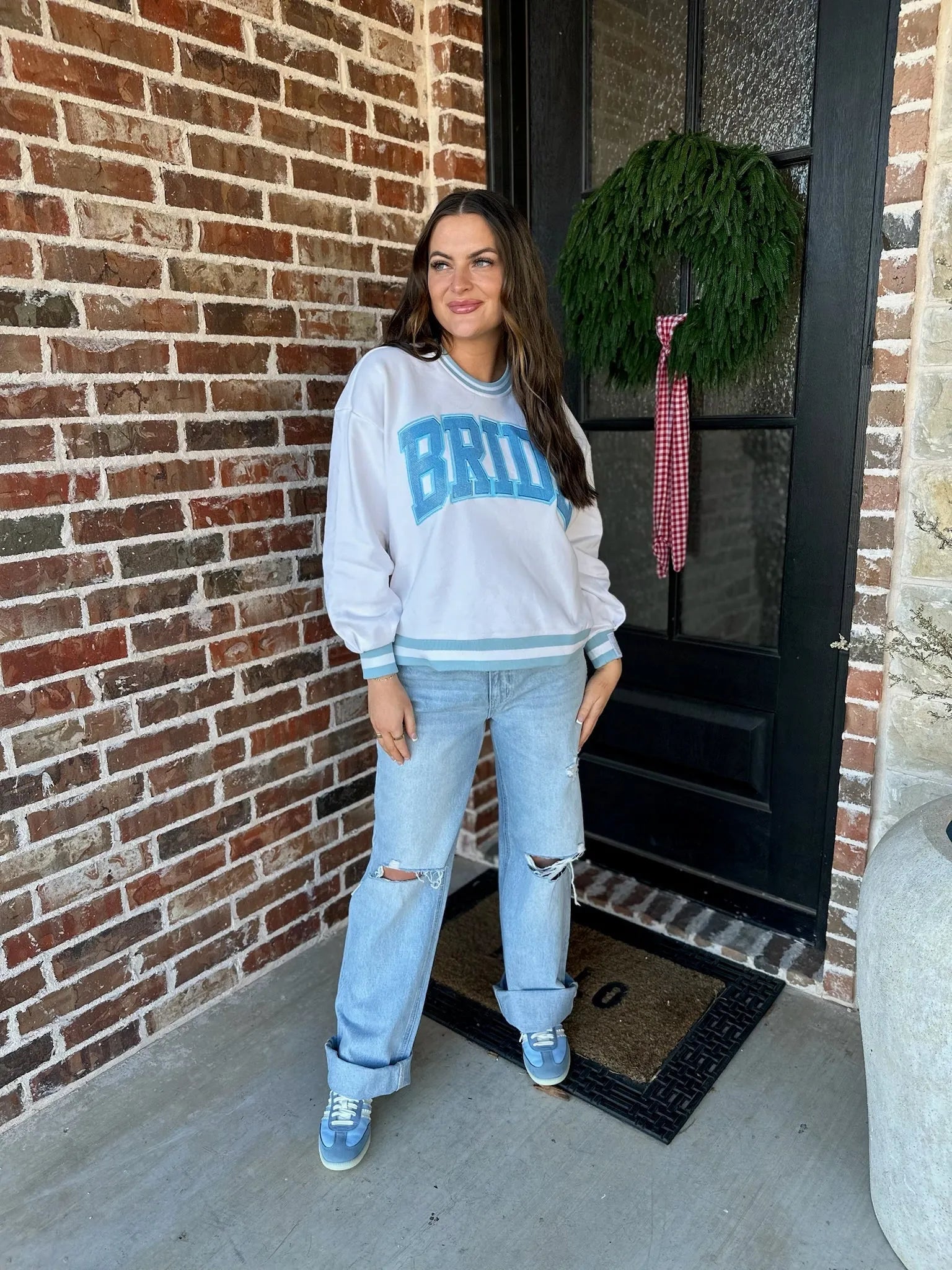 White sweatshirt with blue "BRIDE" lettering, blue trim, paired with distressed light wash jeans and blue sneakers. Model