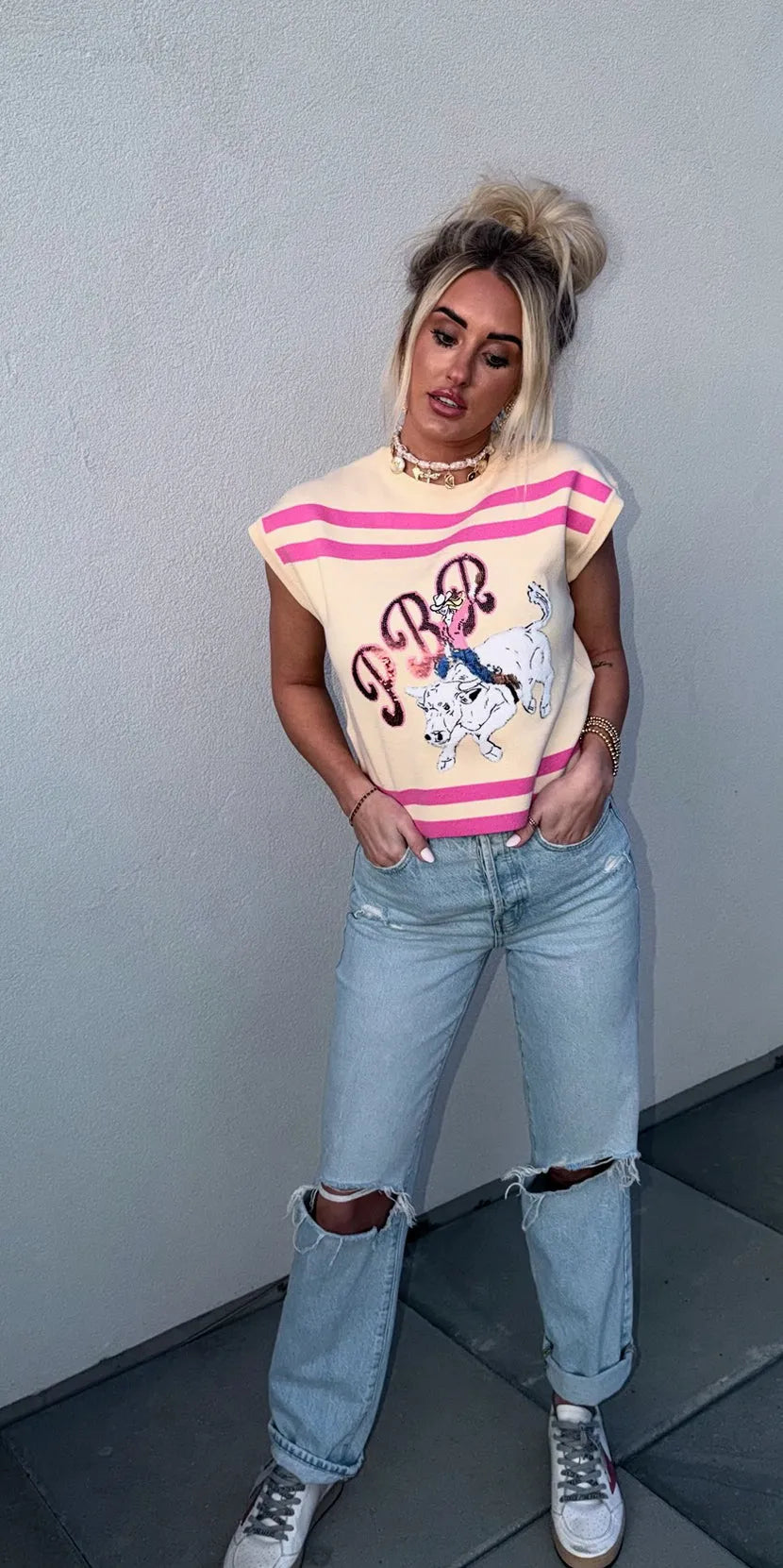 Queen of Sparkles tan knit tank top with pink stripes and a PBR cowgirl graphic. Woman wearing ripped light wash jeans and