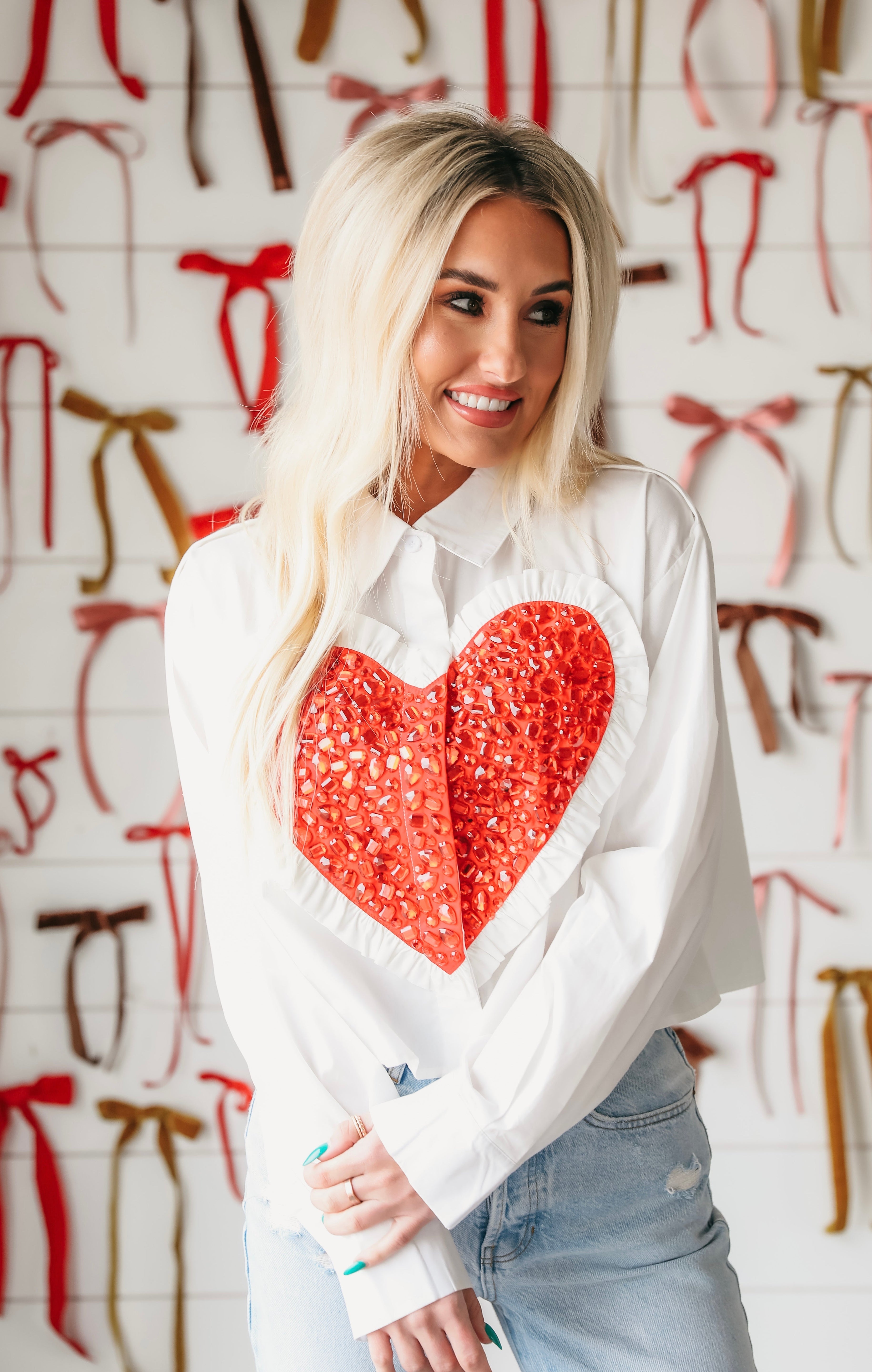 White button-down shirt with a large, ruffled red sequin heart applique on the chest. Model wears light wash jeans