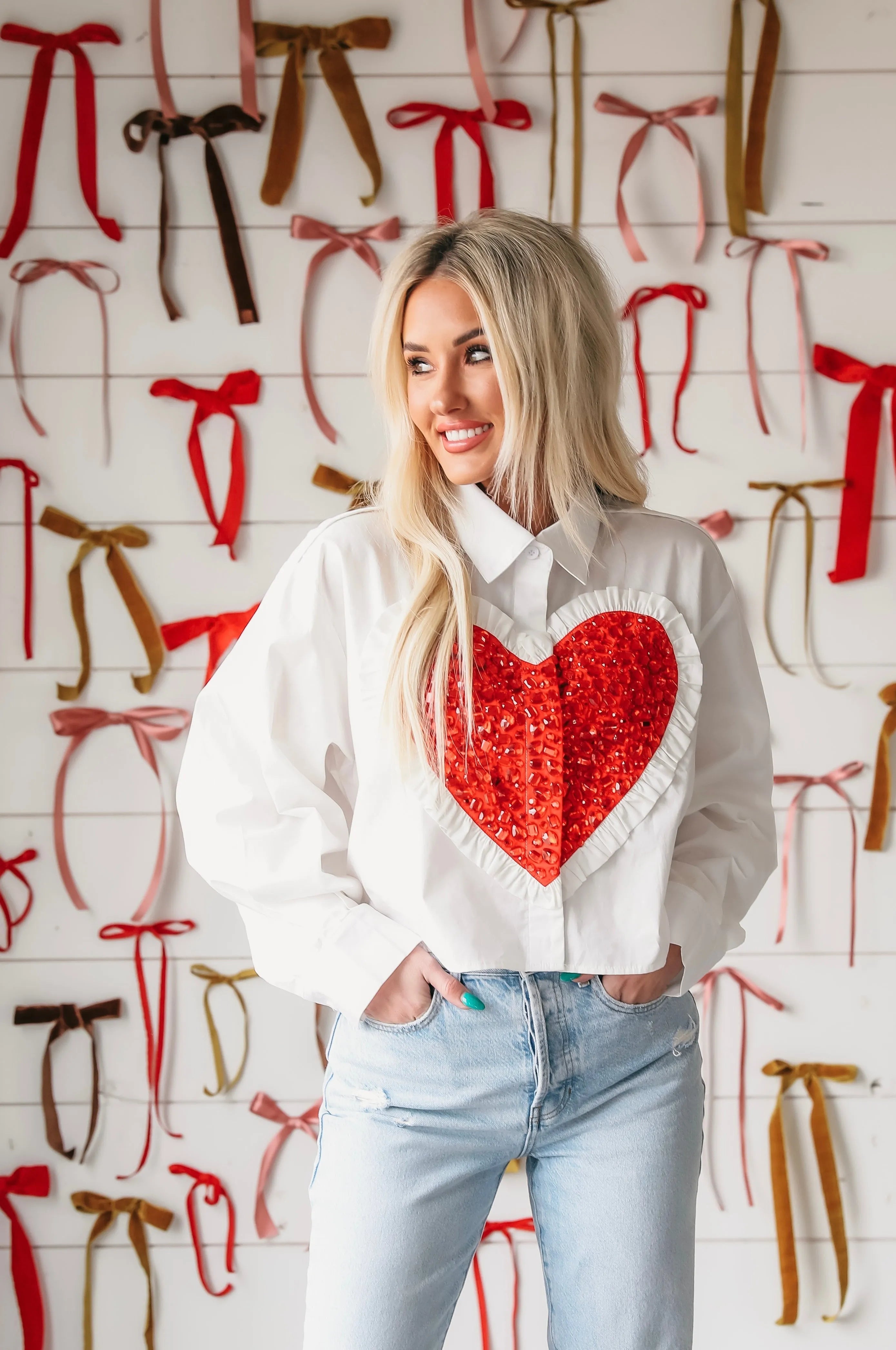 Woman wearing a white button-down shirt with a large, sequined red heart applique on the front and ruffled trim. Light wash