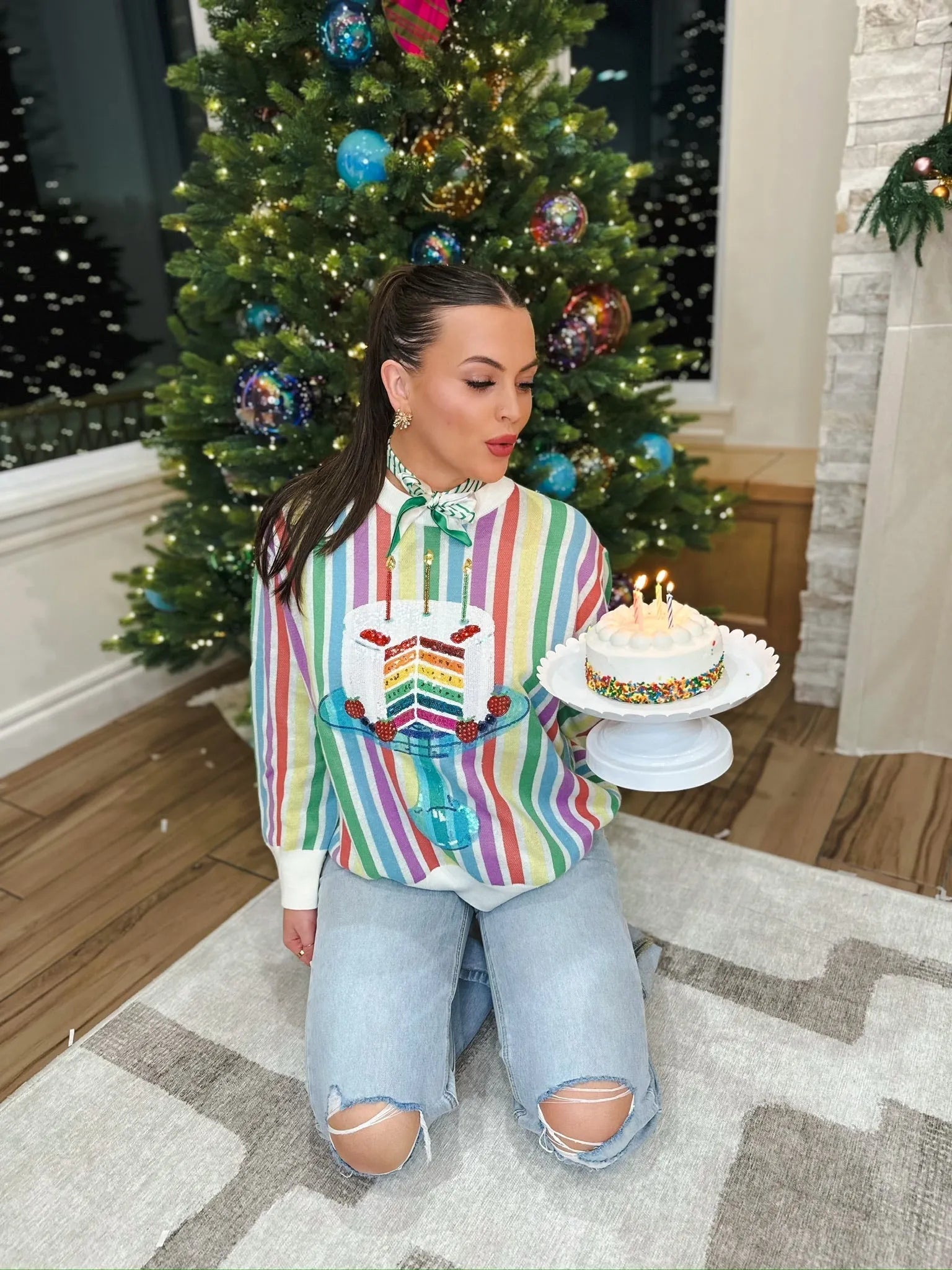 Woman wearing a rainbow striped sweater with a sequined birthday cake graphic, holding a small cake, in front of a