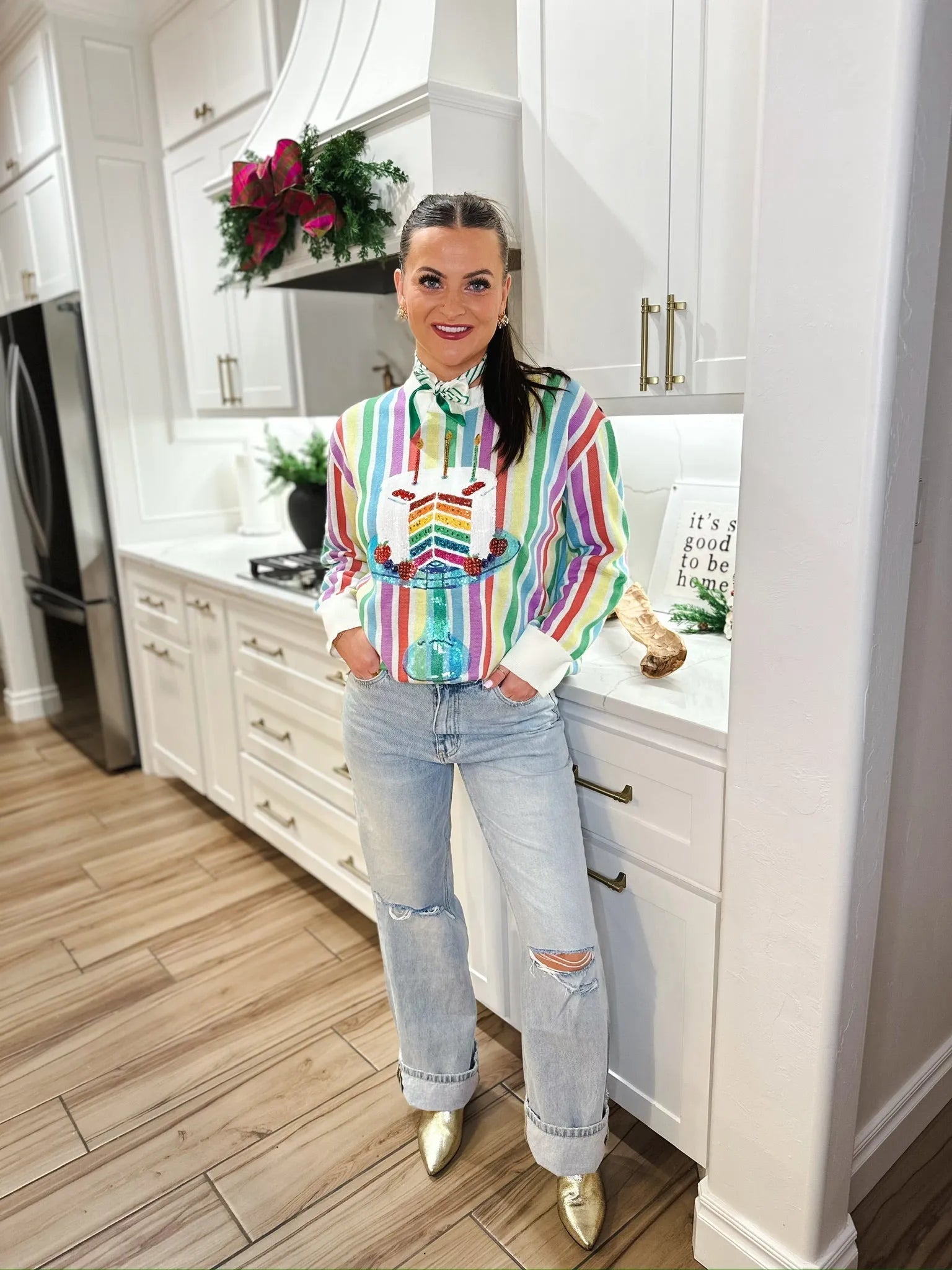 Woman wearing a rainbow stripe sweater with a sequined birthday cake design and light wash ripped jeans.