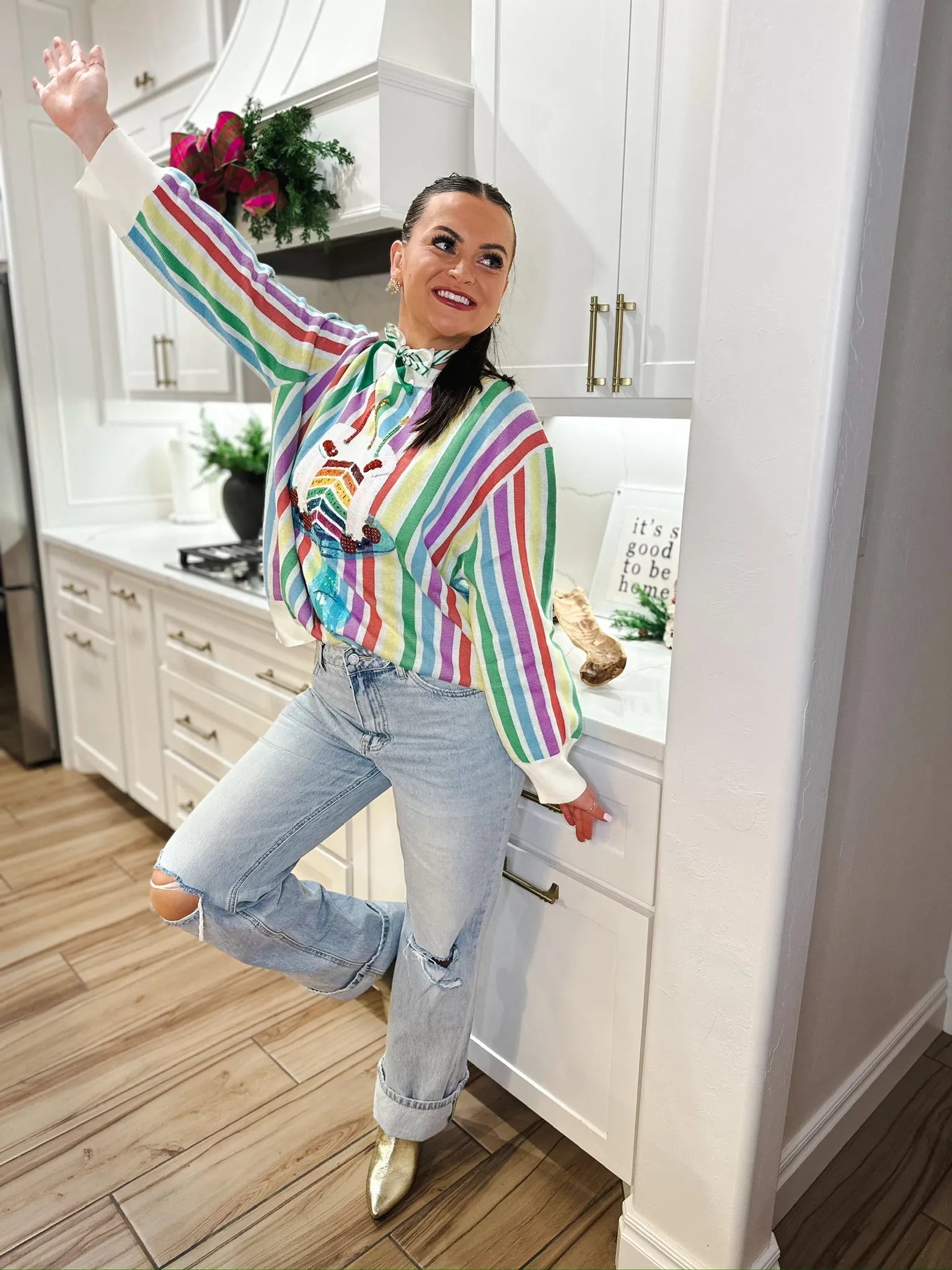 Woman wearing a rainbow stripe sweater with a cake graphic and distressed jeans, posing in a kitchen.