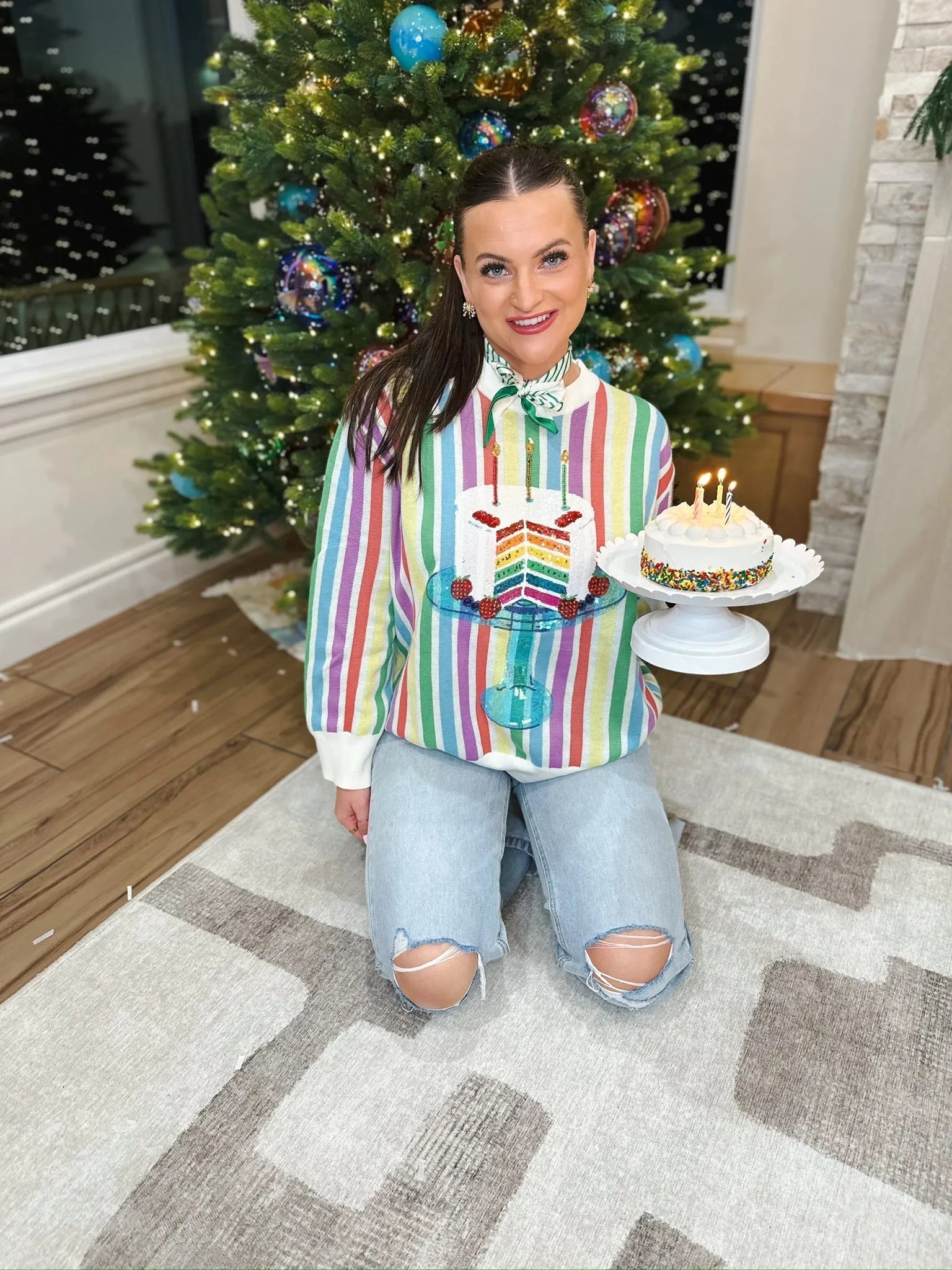 Woman wearing a rainbow stripe sweater with a sequin birthday cake graphic, holding a small cake, in front of a decorated