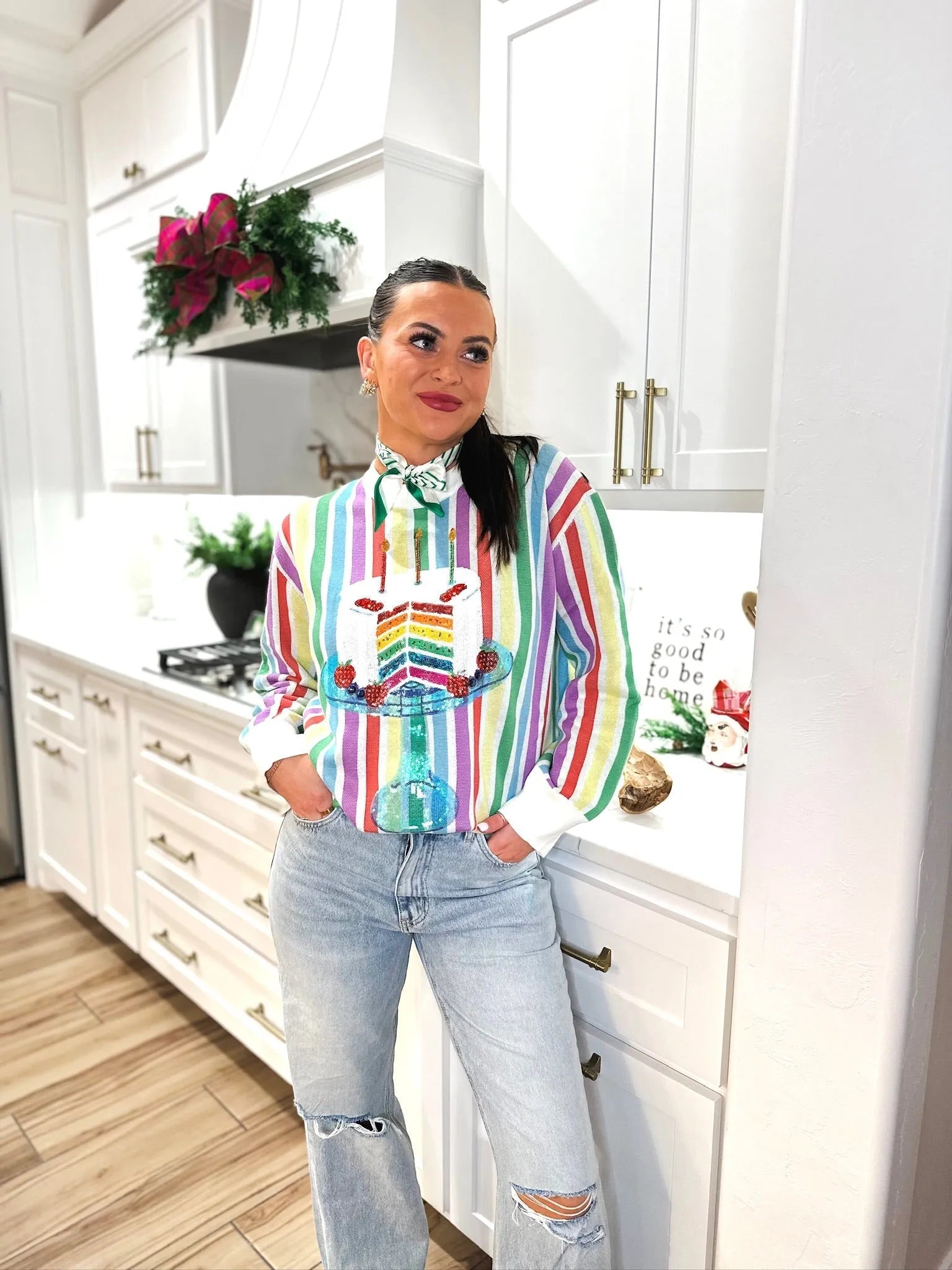 Woman wearing a rainbow striped sweater with a sequined birthday cake graphic. The sweater has long sleeves and a white