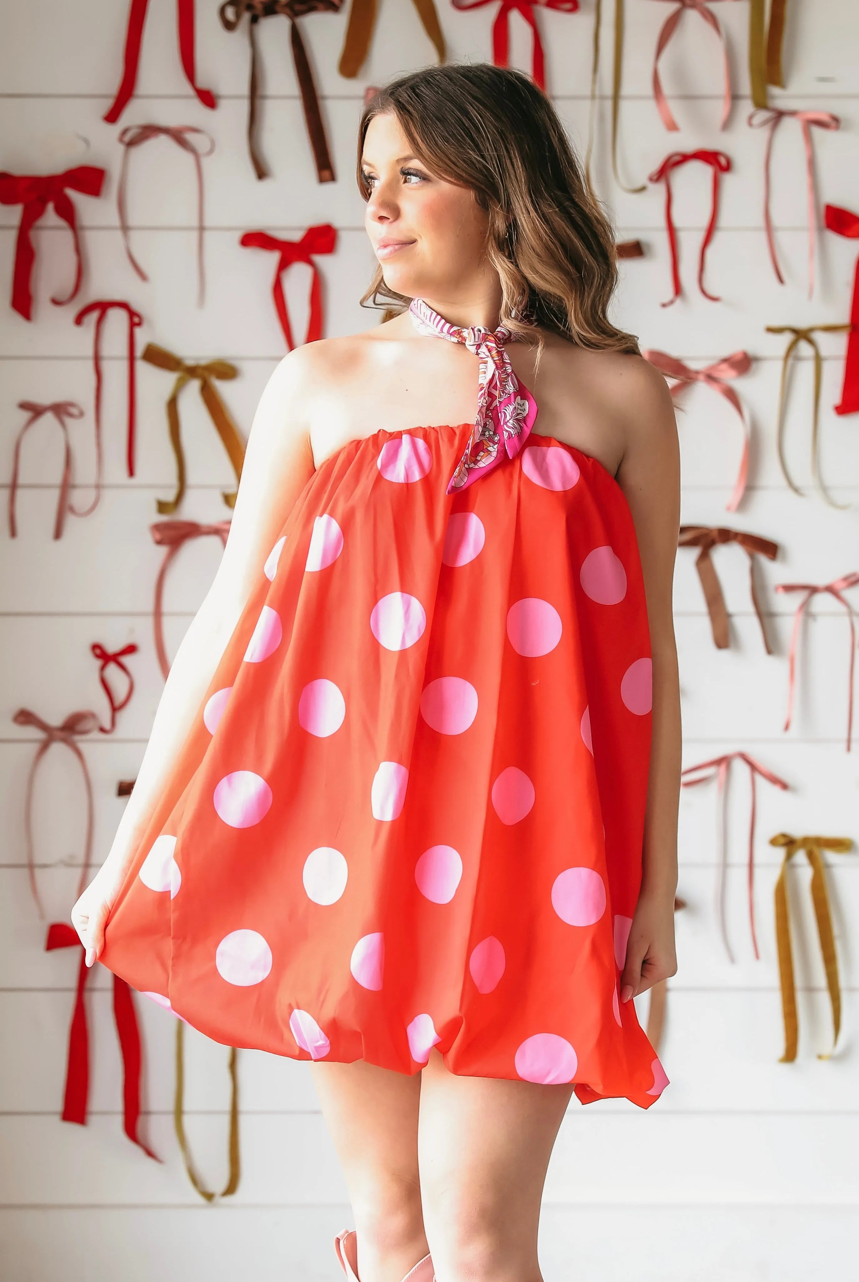 A woman wears a red and pink polka dot strapless bubble dress, paired with pink cowboy boots. She has a scarf tied around