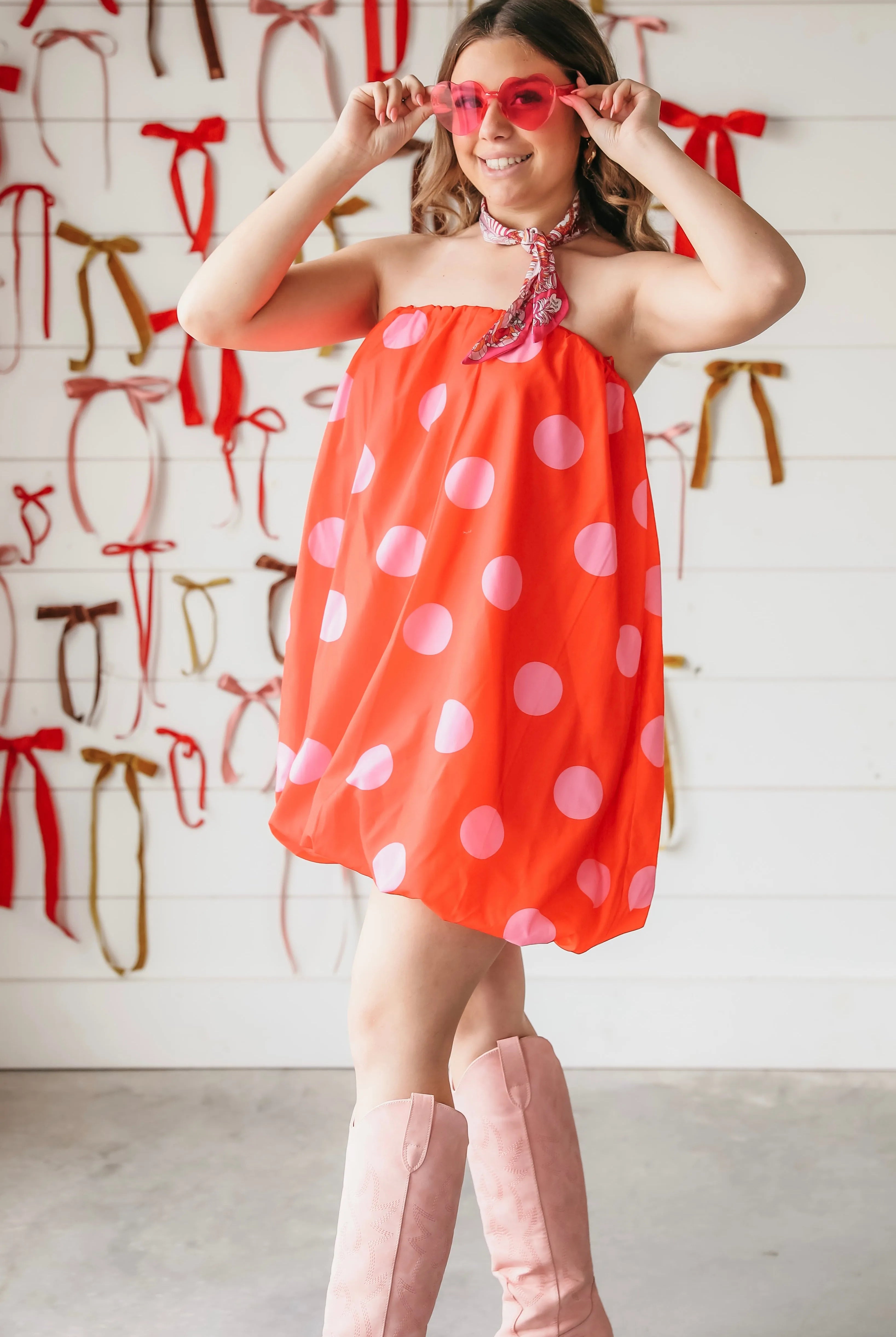 Woman in red and pink polka dot strapless bubble dress, pink heart sunglasses, and pink cowboy boots.