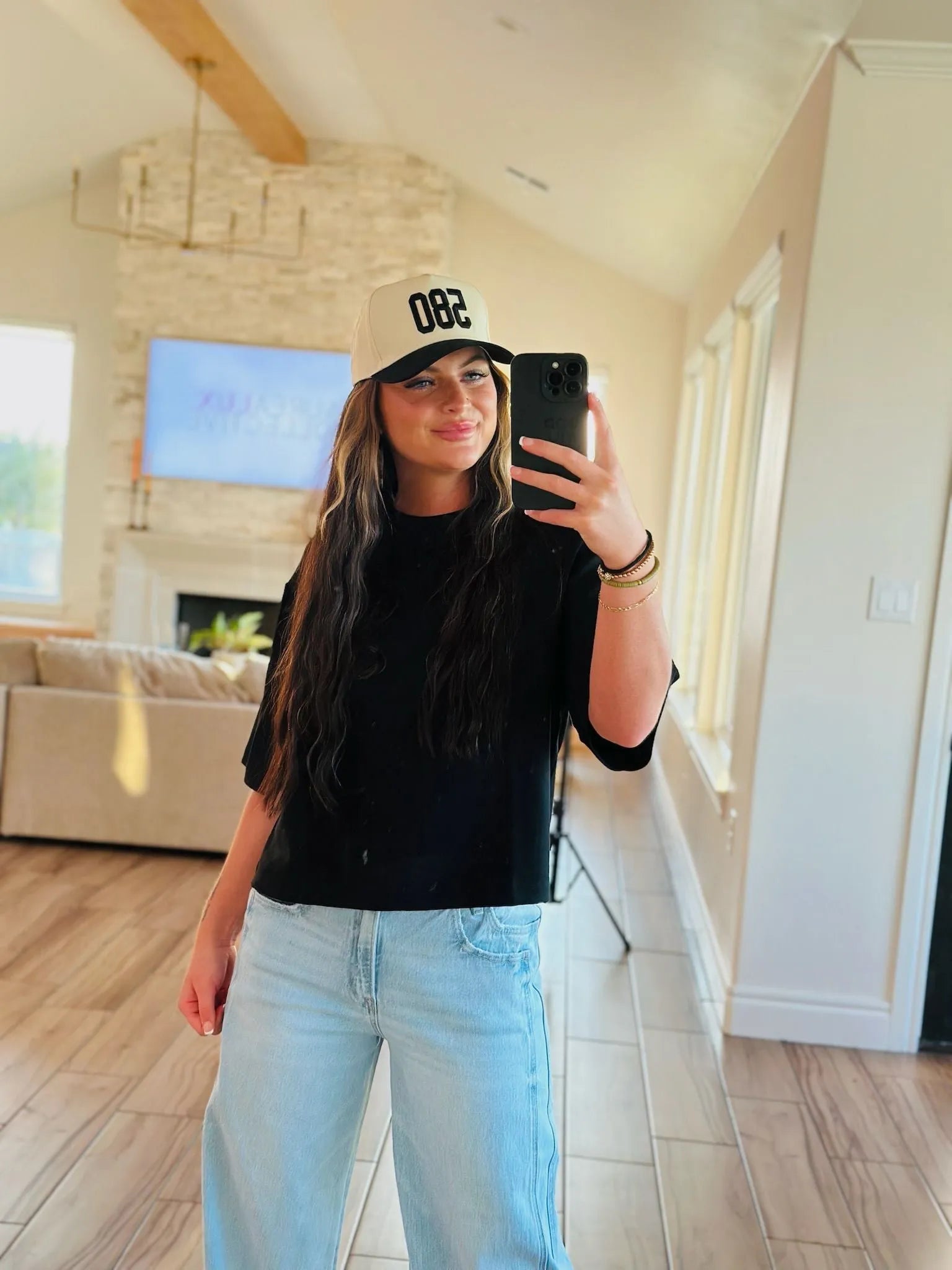 REPRESENT 580 trucker hat in black and beige with custom area code "082" on front. Woman wearing hat, black t-shirt, and