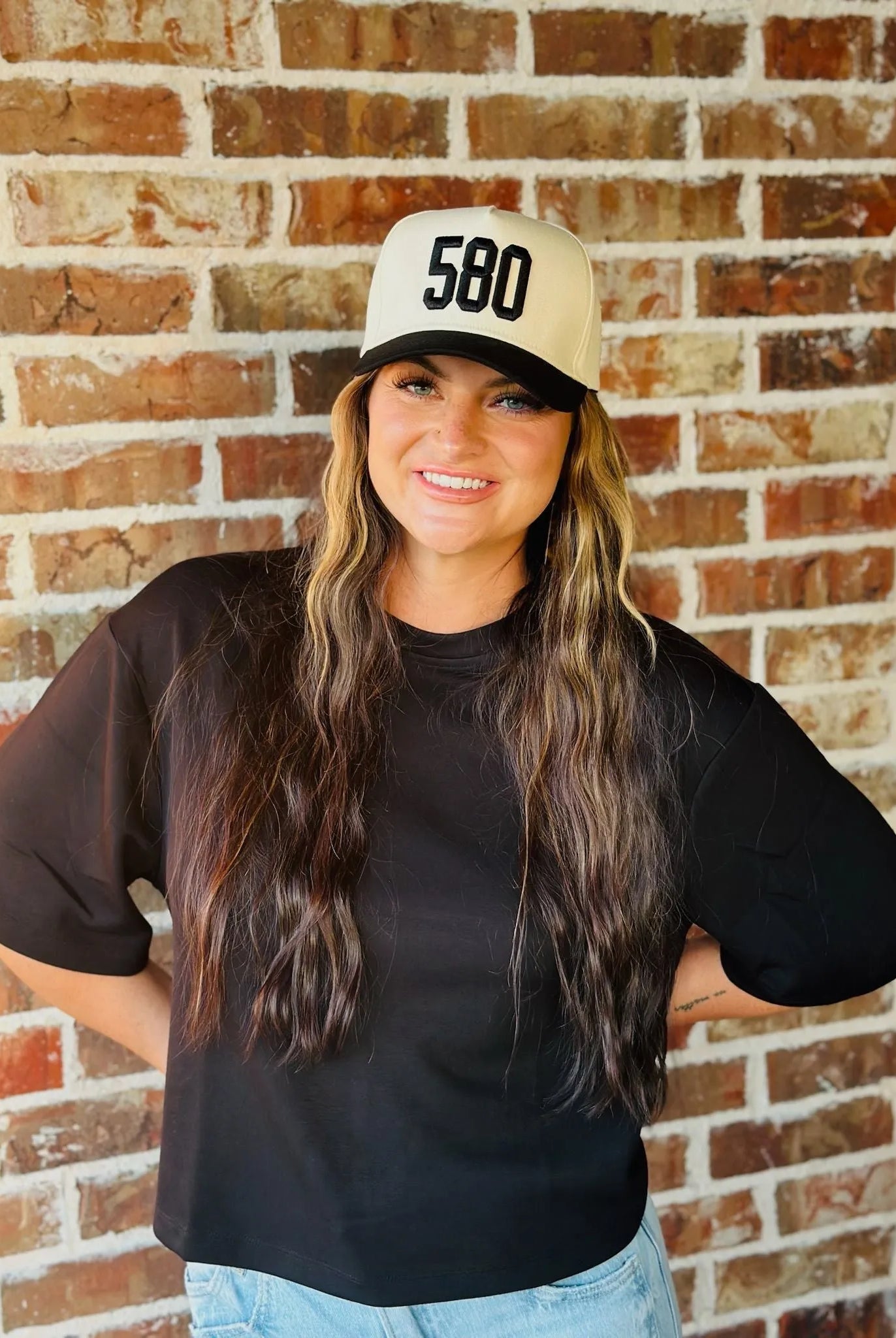 Woman wearing a black REPRESENT 580 custom area code trucker hat with a cream-colored front and black brim.