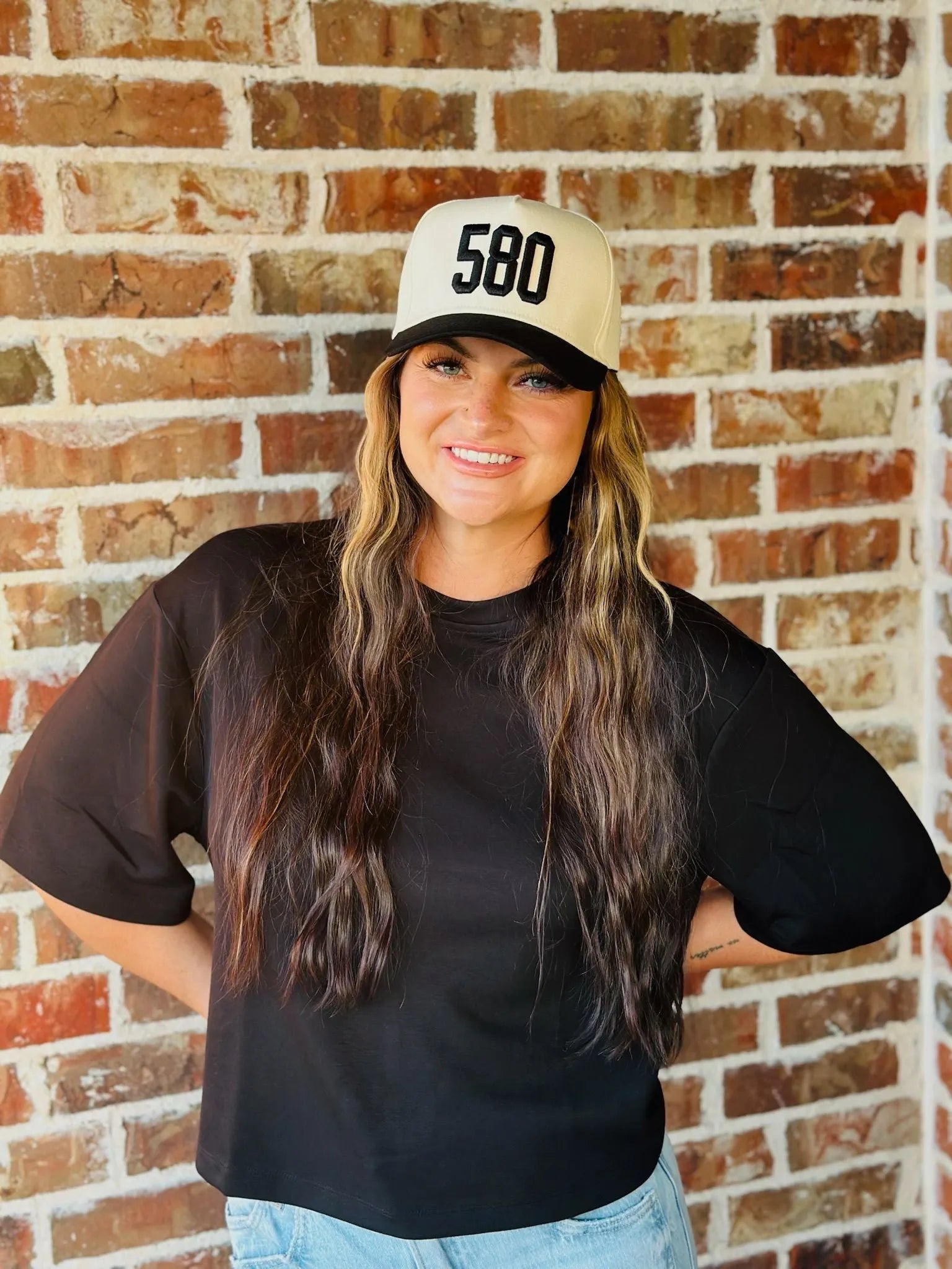 Woman wearing a black REPRESENT 580 custom area code trucker hat with a cream-colored front and black brim.