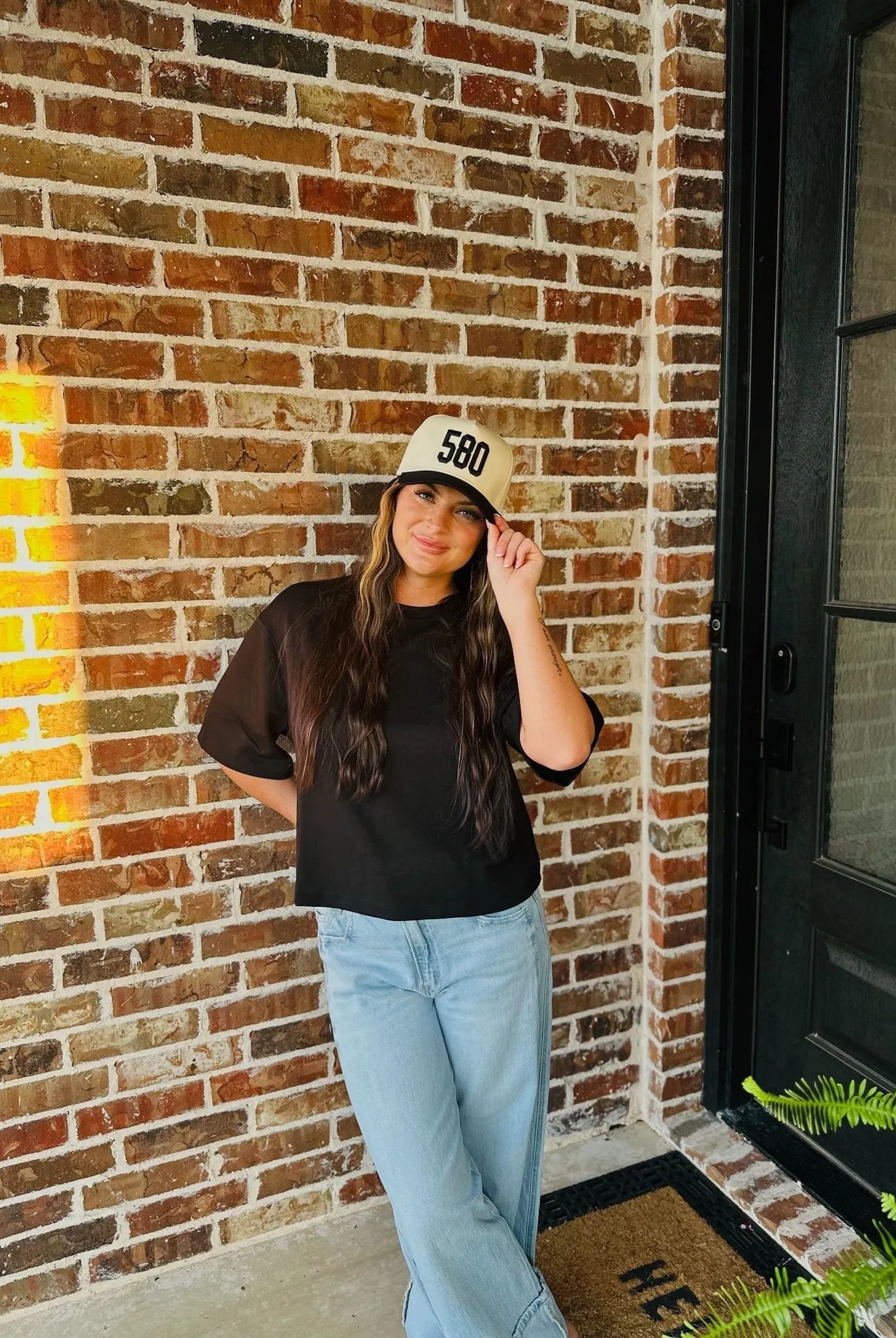 Woman wearing a black REPRESENT 580 custom area code trucker hat on a brick background.