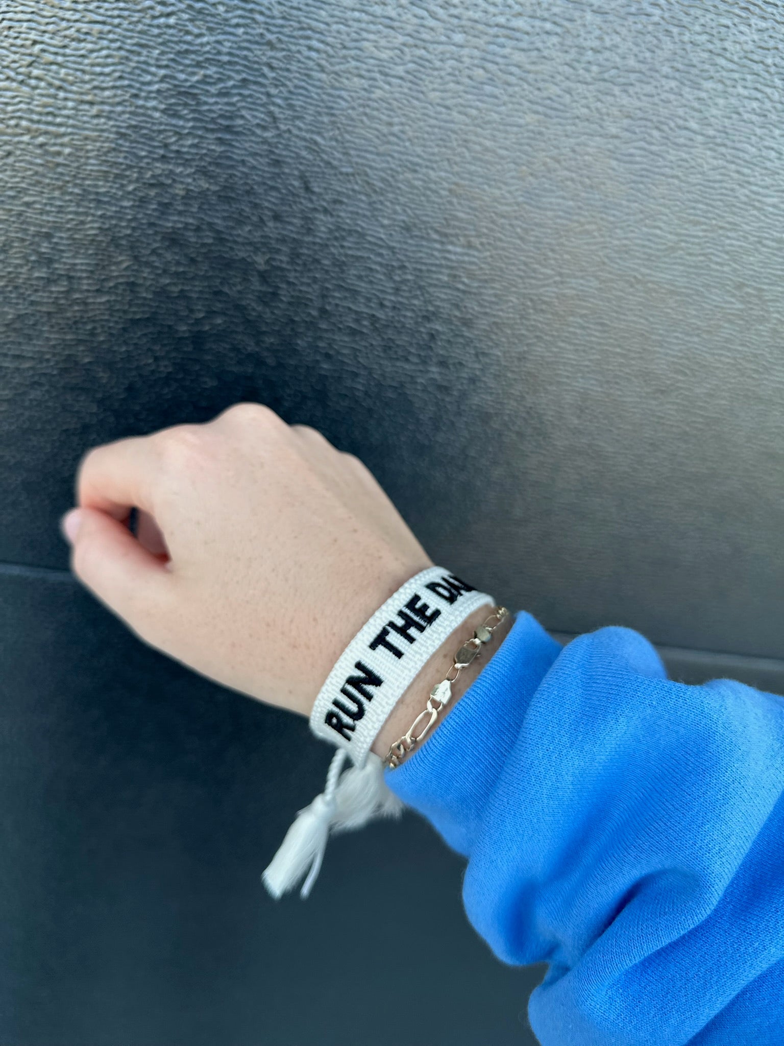 White woven bracelet with black embroidered text 'RUN THE DA' and a gold chain bracelet stacked below it on a wrist.