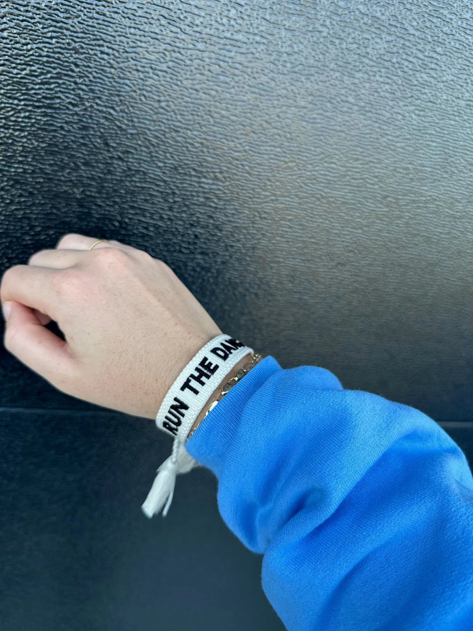 White woven bracelet with black embroidered text 'RUN THE DA...' on a wrist, over a blue sleeve.