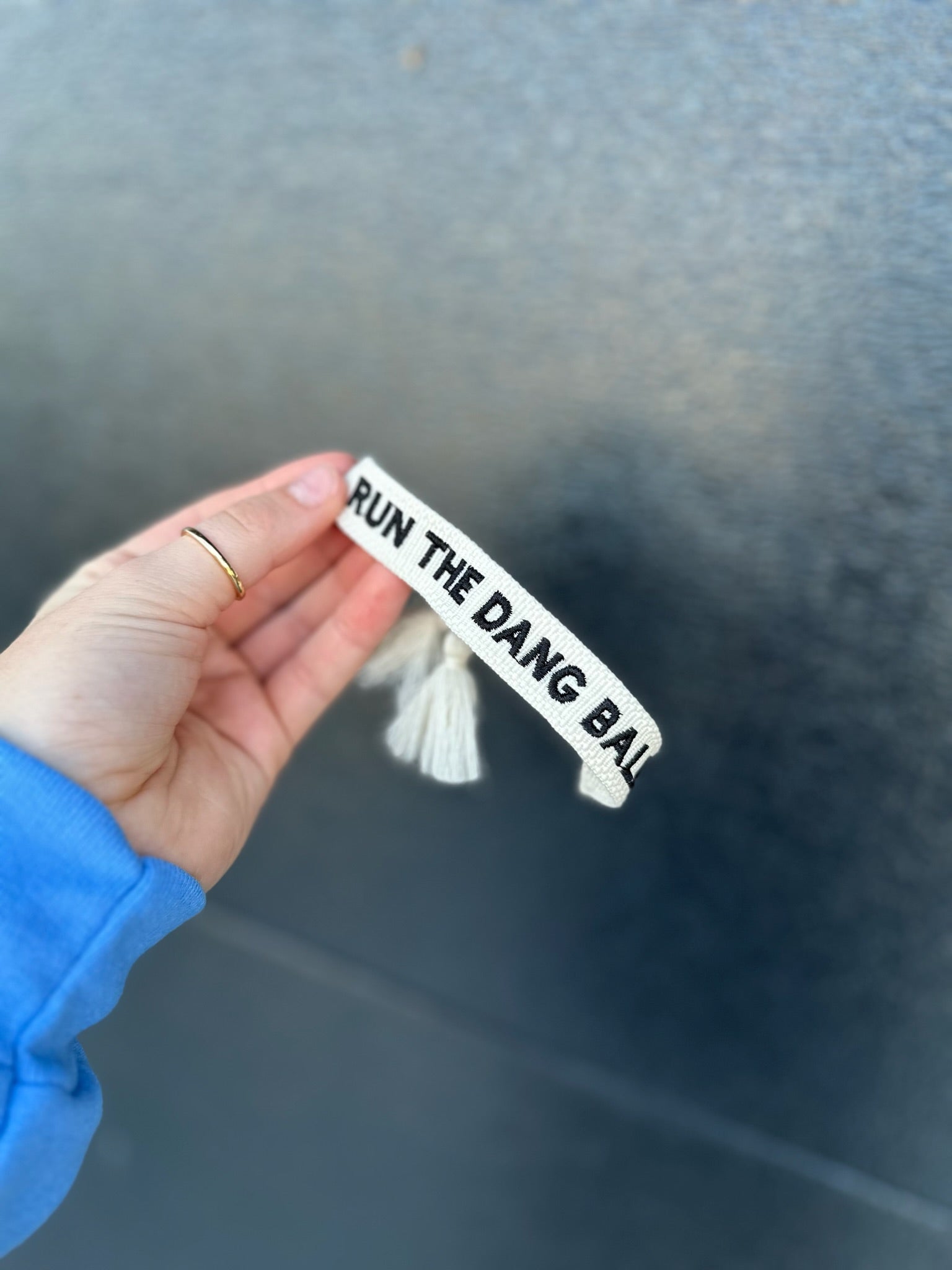 White embroidered bracelet with black text 'RUN THE DANG BAL' and two white tassels, held by a hand with a gold ring.