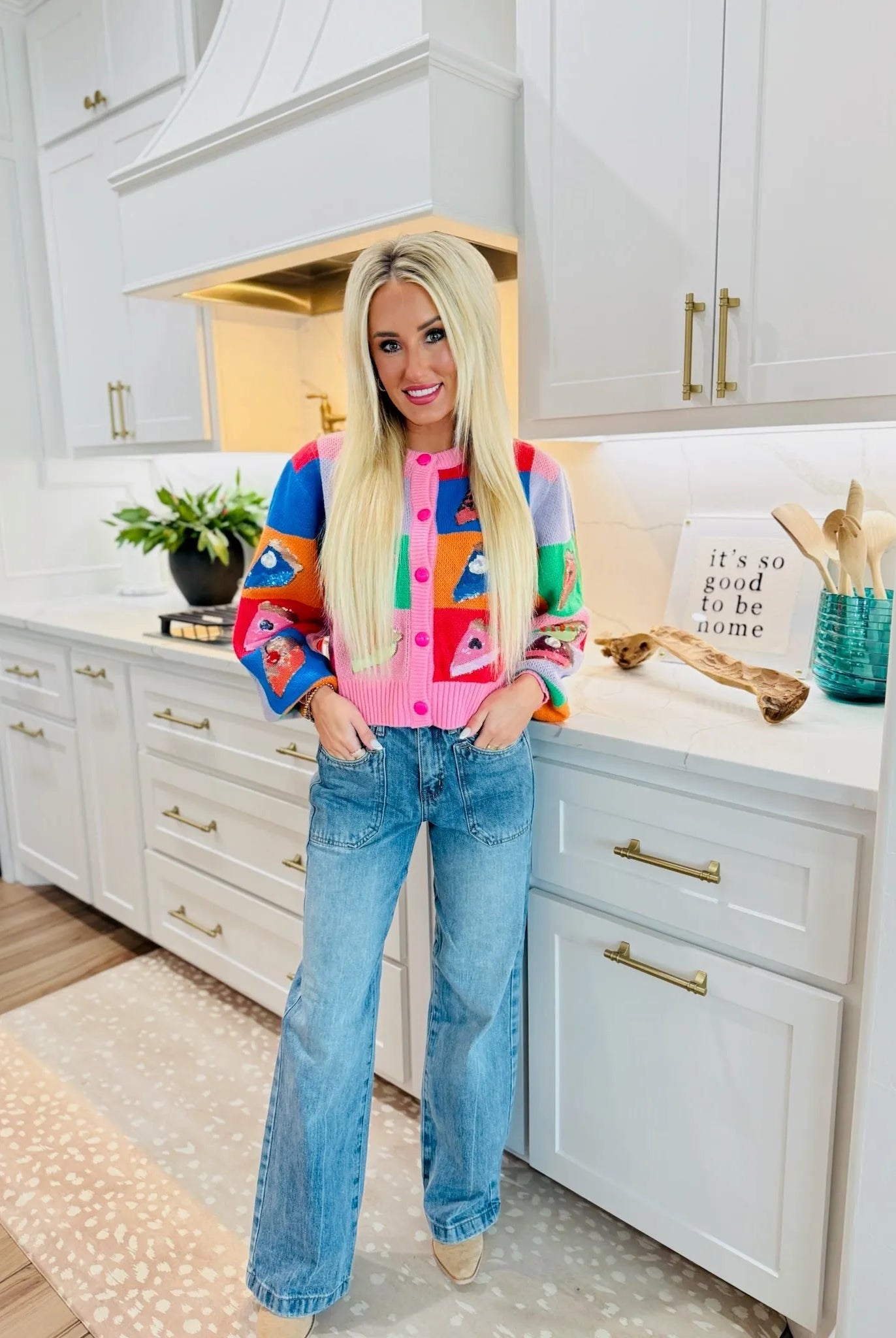 Woman wearing SAIGE high-rise double-pocket wide-leg jeans and a colorful fruit-print cardigan, standing in a kitchen.