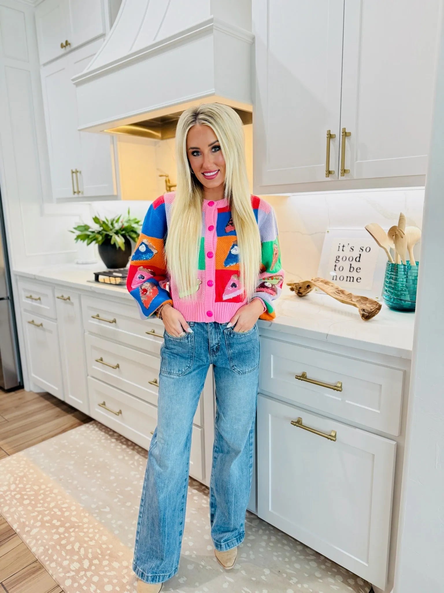Woman wearing SAIGE high-rise double-pocket wide-leg jeans and a colorful fruit-print cardigan, standing in a kitchen.