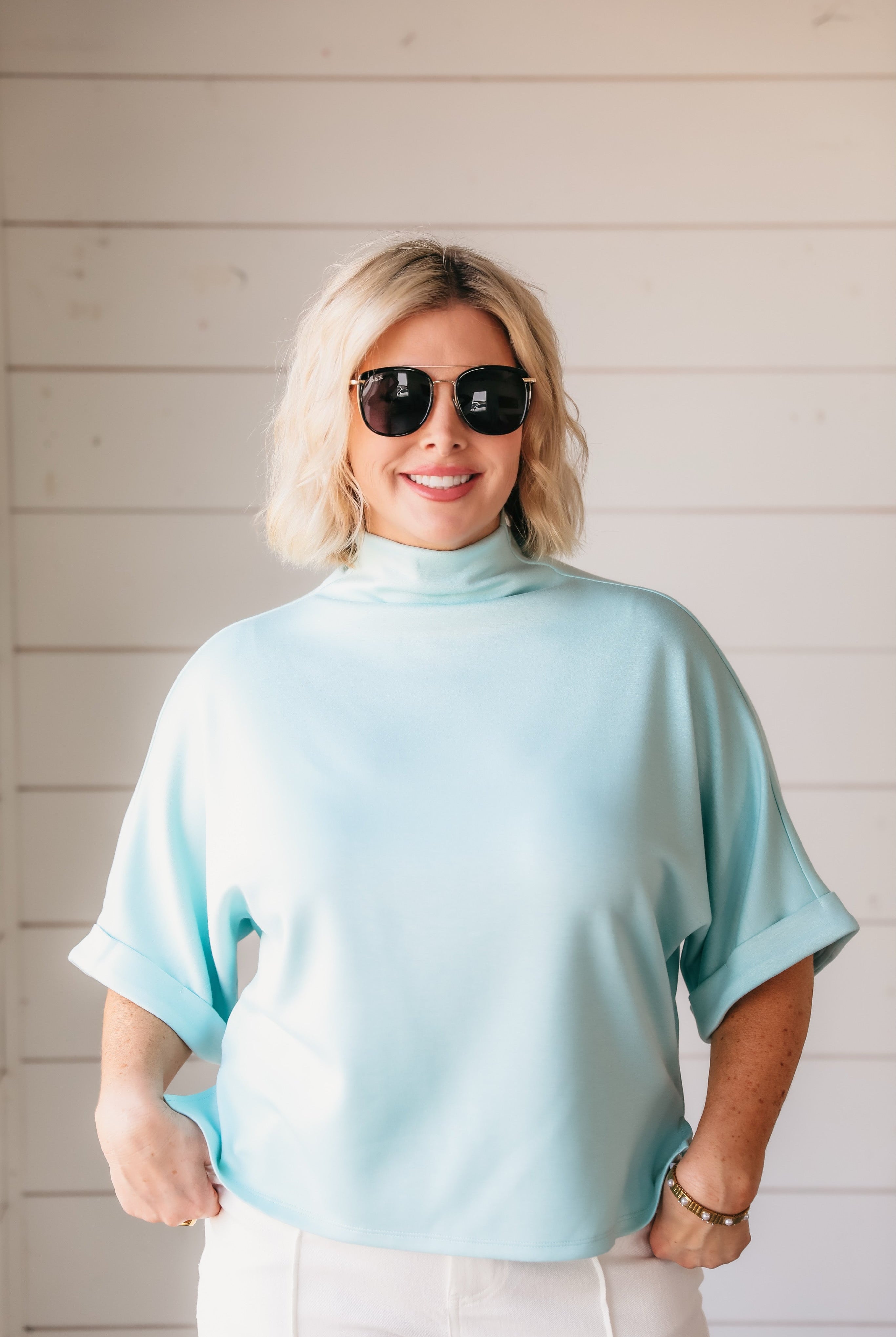Woman wearing a seafoam soft knit spring top with a mock neck and cuffed short sleeves. She wears sunglasses and white pants.