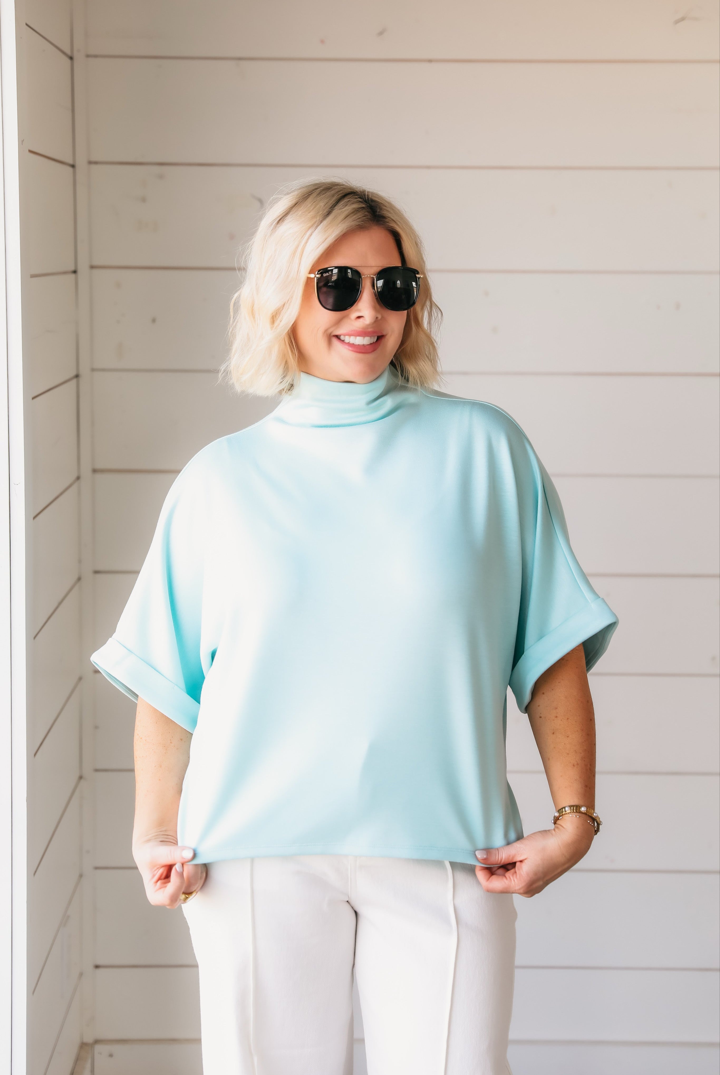 Woman wearing a sea foam soft knit spring top with a mock neck and cuffed short sleeves, paired with white pants and