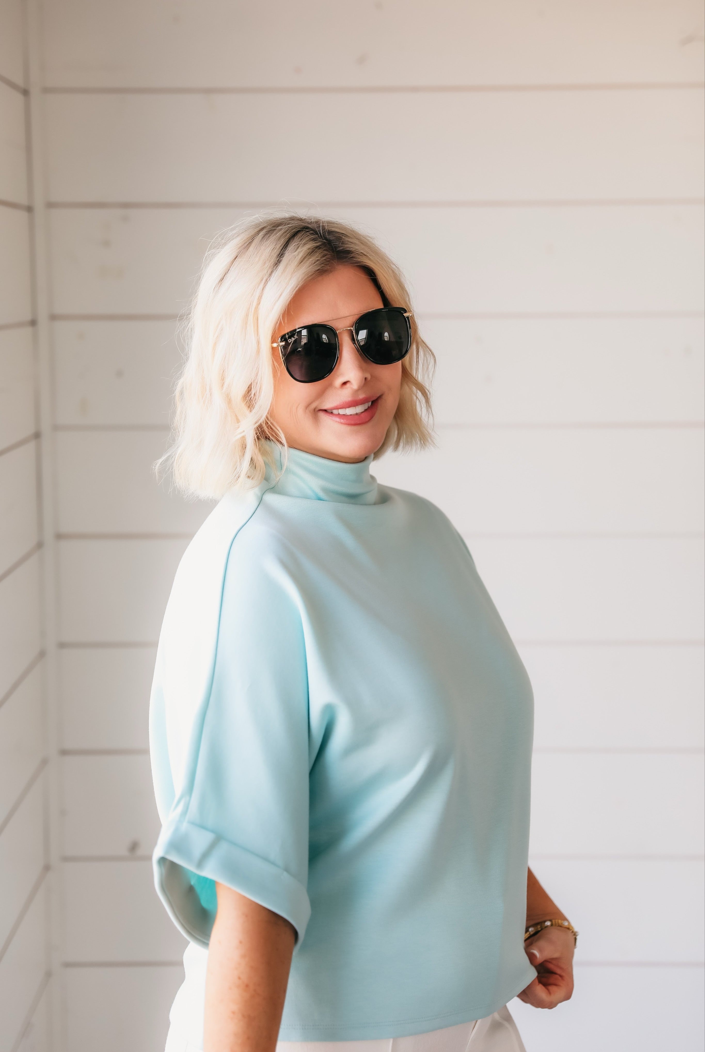 Woman wearing a sea foam green soft knit mock neck top with cuffed sleeves. She has blonde hair and is wearing sunglasses.