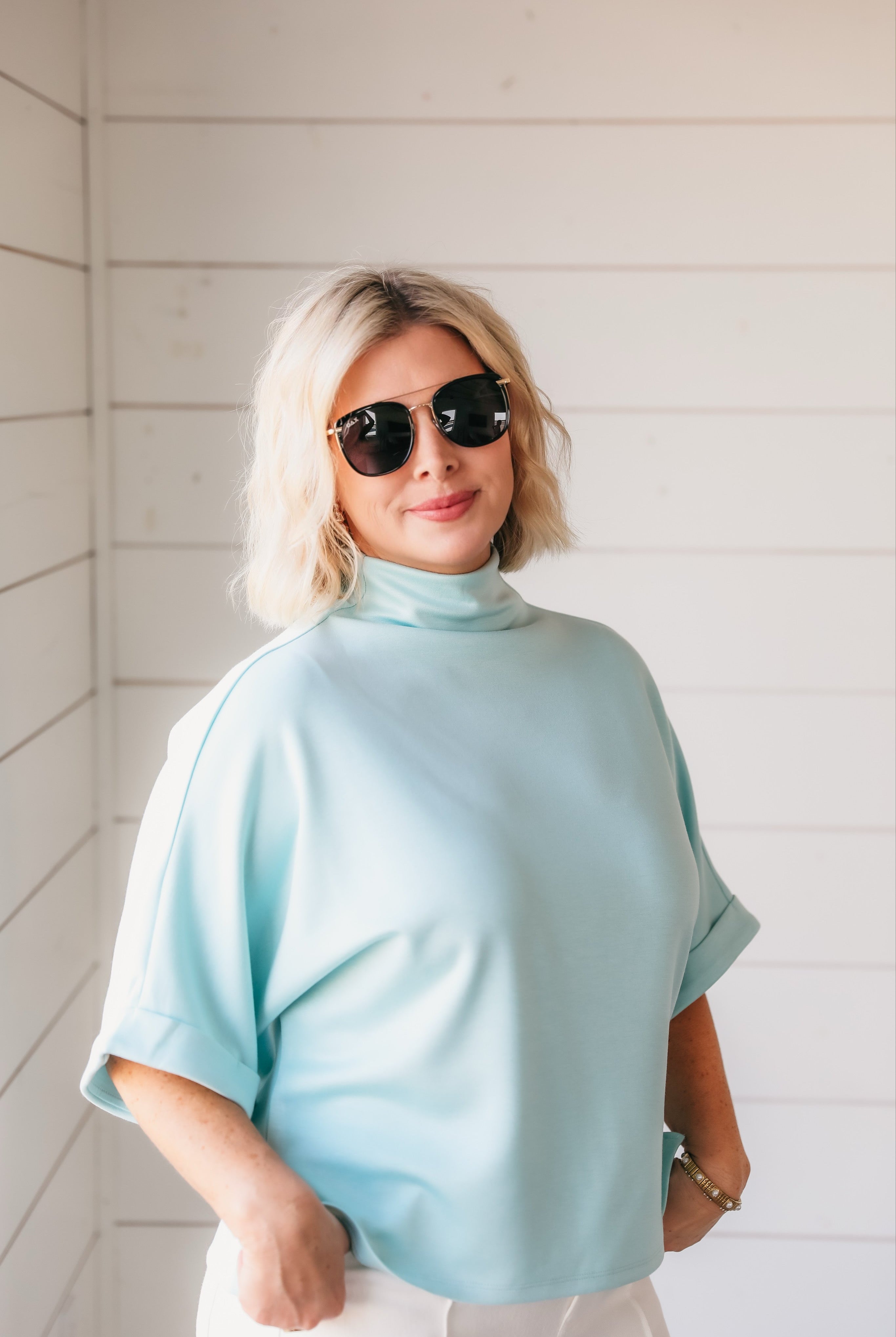 Woman wearing a sea foam soft knit spring top with a turtleneck, cuffed short sleeves, and sunglasses.