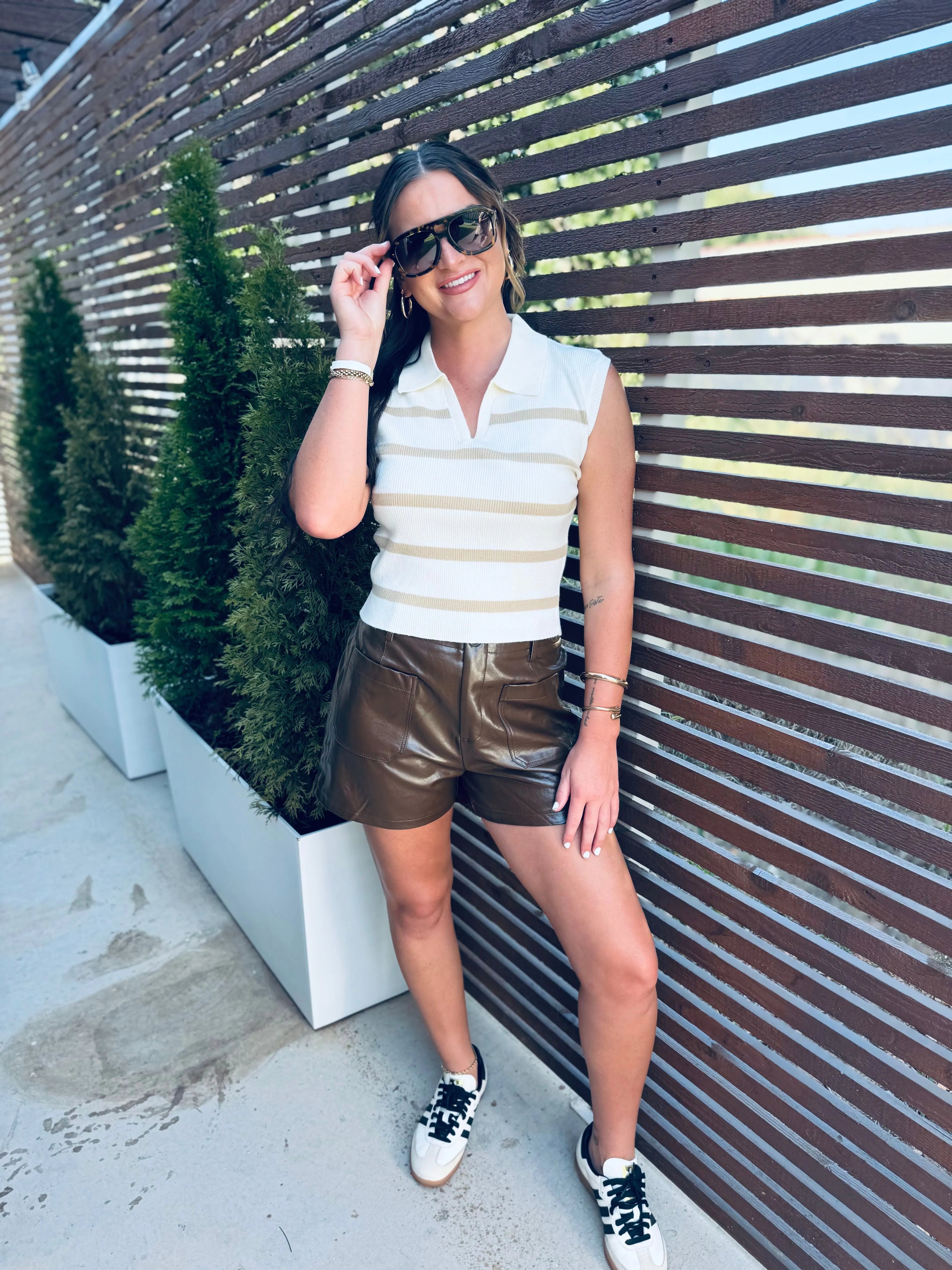 Woman wearing a white and tan striped knit polo vest, brown faux leather shorts, and white sneakers. She is wearing